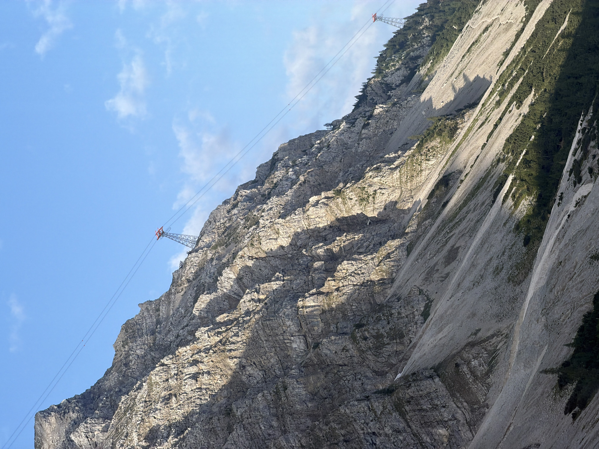 The image depicts a steep, rocky mountain slope with a cable car system traversing it. The mountain is characterized by its rugged texture, with visible layers of rock and patches of sparse vegetation. Sunlight casts dramatic shadows across the slope, highlighting the contours of the terrain.
In the upper portion of the image, a red and silver cable car tower is visible, with cables extending diagonally across the sky. Further up the slope, another similar tower can be seen. The sky is a clear blue, with a few scattered white clouds.
The lower part of the mountain is covered in scree and some scattered evergreen trees. A narrow, winding path is visible on the slope, suggesting a hiking trail. There are no people clearly identifiable in the image.
This scene is characteristic of the Bavarian Alps region, and specifically, the area around Grainau, Germany, which is known for its mountainous terrain and cable car systems that provide access to higher elevations for tourism and recreation.