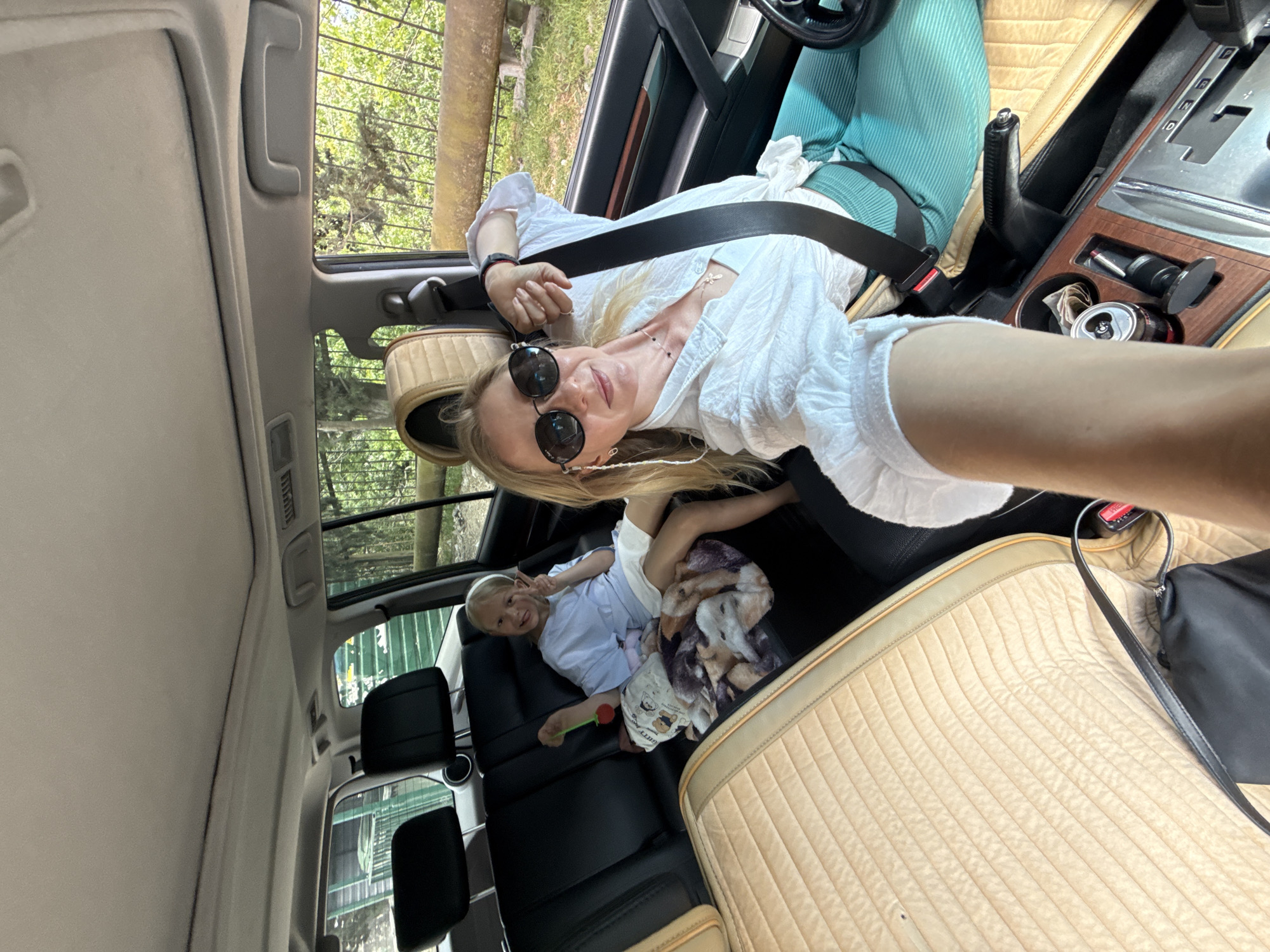 The image is a selfie taken inside a vehicle, possibly a car. The photographer is a blonde woman wearing sunglasses and a white top, with a seatbelt across her chest. She is looking at the camera. In the back seat, there is a child lying down on a blanket, looking towards the camera and holding up a peace sign with their fingers. The interior of the vehicle has light-colored, quilted seats, and the dashboard area is visible. Outside the car windows, there are trees and foliage, suggesting the vehicle is parked in a wooded area. The location or city is not discernible from the image.