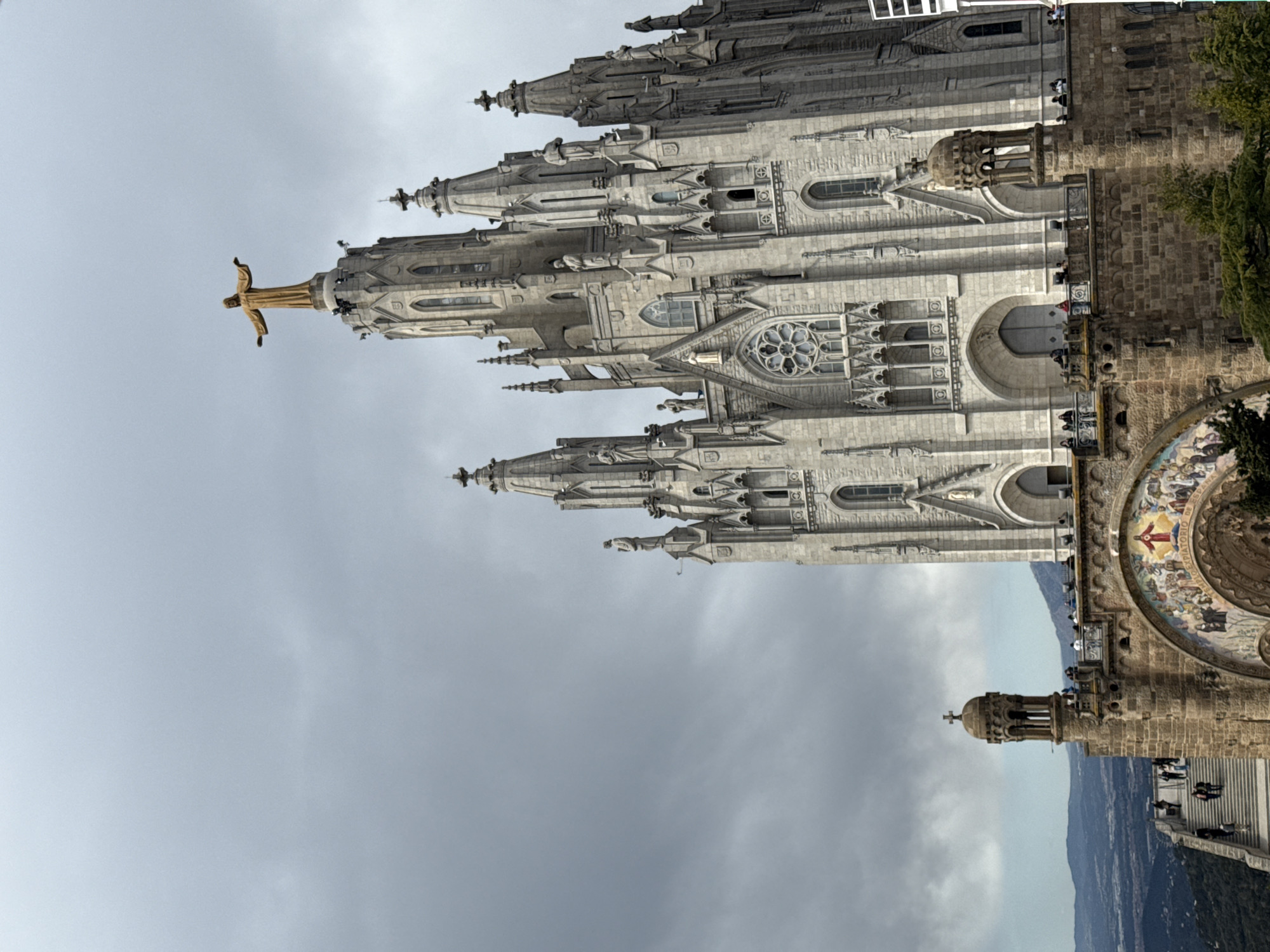 The image shows the Sagrat Cor temple at the summit of Mount Tibidabo, in Barcelona, Spain. The temple is a large, predominantly white or light-gray stone structure with Gothic architectural features. A golden statue of Christ with outstretched arms stands atop the main tower. People can be seen on the side of the building, some appearing on balconies or terraces. A section of the building features a mosaic and additional religious imagery. The environment includes a cloudy sky and a glimpse of the surrounding landscape from the mountaintop.