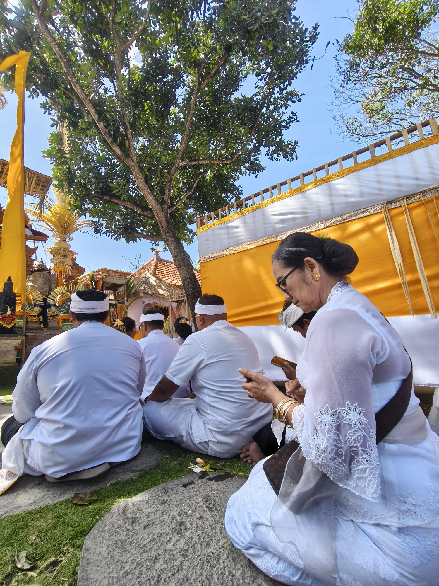 In Denpasar, Indonesia, people are participating in a religious ceremony. The image captures a group of individuals, primarily men, dressed in traditional white attire, seated on the ground under a tree. They are facing a structure that appears to be a temple or a religious shrine, adorned with yellow and white fabric, intricate carvings, and offerings. A woman in a white outfit with glasses and elaborate jewelry is also present, holding a smartphone. The scene is set outdoors, with lush greenery and a clear blue sky overhead. The overall ambiance suggests a solemn and respectful gathering.
