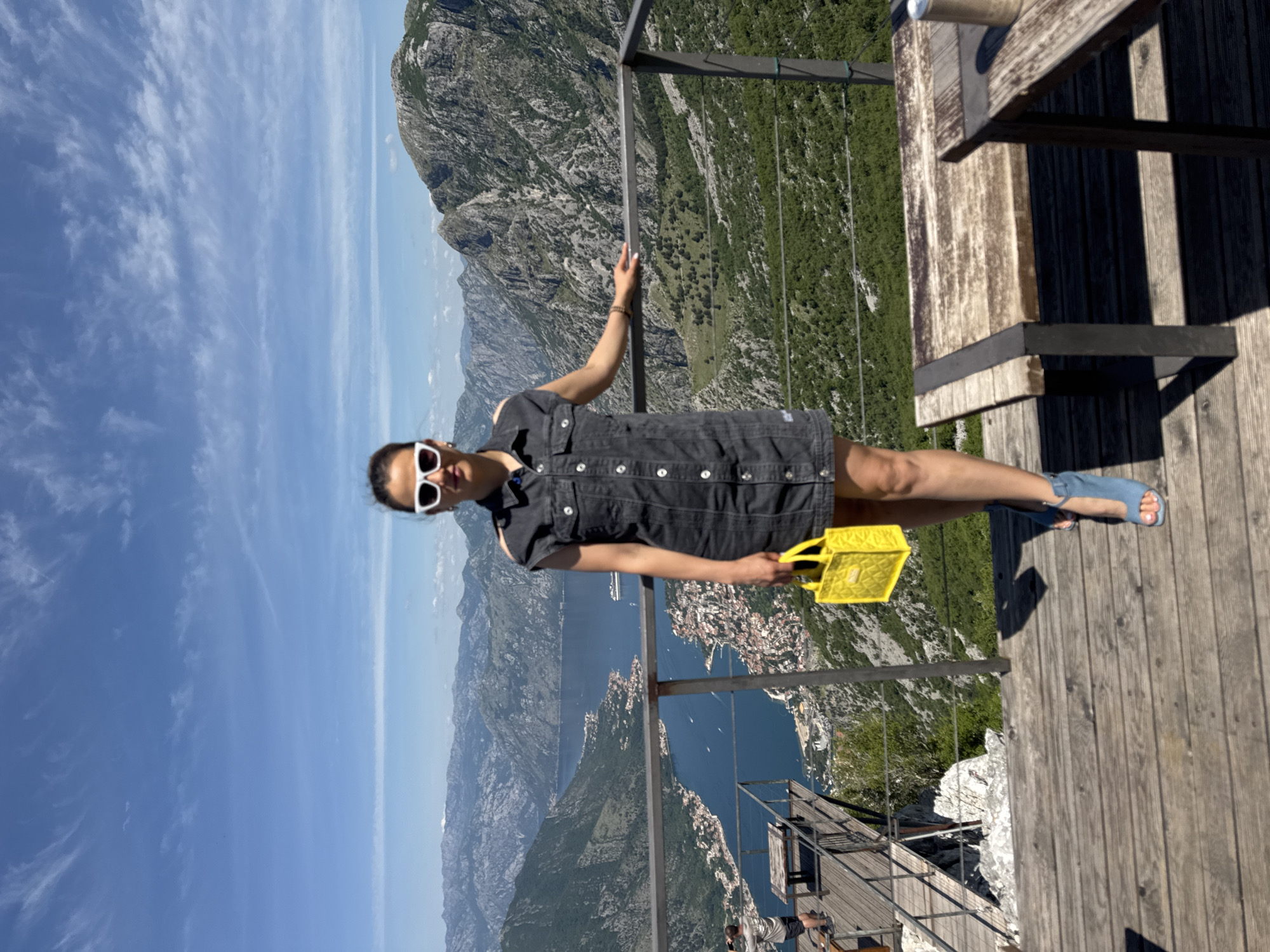 A woman stands on a wooden platform in Žanjev Do, Montenegro, against a backdrop of mountains, a body of water, and the city below. She is wearing a black dress, white sunglasses, and blue sandals. She holds a yellow bag. A metal railing is present, and a wooden table and benches are visible on the platform.