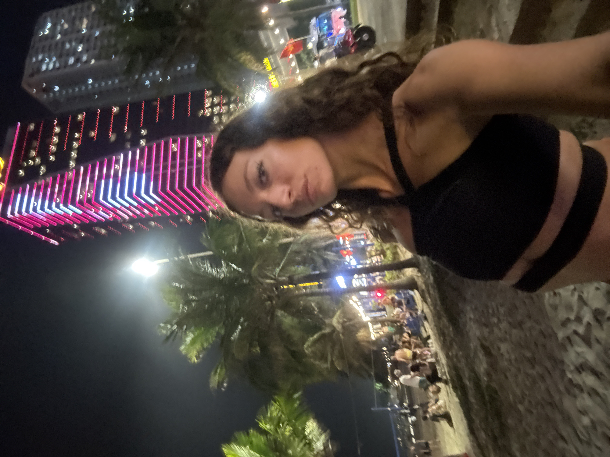 A woman with curly brown hair and wearing a black sports bra is photographed outdoors at night in Đà Nẵng, Vietnam. She is looking at the camera. Palm trees and a bright street light are behind her. In the background are lit buildings, including one with pink and blue neon lights in a patterned design. There is a crowd of people on the right side of the frame. The ground appears to be paved. A building with white lights is also visible.