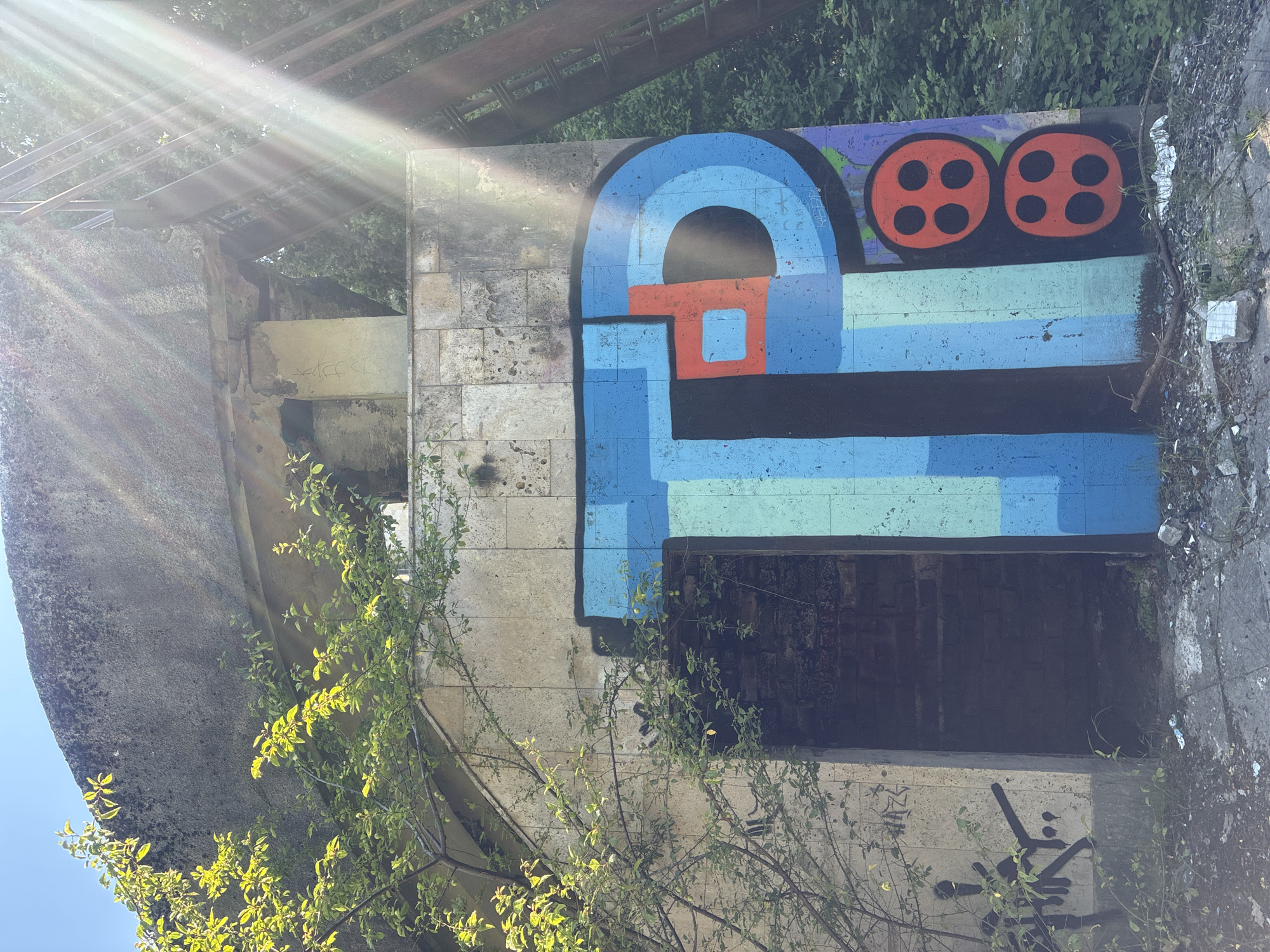 The image, taken in Sochi, Russia, features a vibrant graffiti artwork on a textured concrete wall, illuminated by bright sunlight. The central graffiti piece is a large, stylized design primarily in shades of blue and black, with an orange rectangular element integrated into an arched section. Above this, two bold red circles, each with four black dots, are painted on a purple and green background, possibly depicting film reels or abstract eyes.
The scene appears to be an outdoor area with robust, aged concrete structures. To the left, a massive, curved concrete form is partially obscured by bright green foliage. The wall where the main graffiti is located is composed of stone-like blocks. Above and behind this wall, a dense canopy of green leaves is visible, along with some metal beams, suggesting an overhead structure such as a bridge or a roof support. Strong sunlight streams from the upper left, creating visible light rays and lens flares.
Below the main graffiti, a dark, rectangular recess or opening is visible in the wall, appearing either boarded up or naturally dark. On the concrete surface directly beneath this opening, a distinct black spray-painted symbol, resembling a stylized 'Y' or an abstract figure, is present, accompanied by some smaller, illegible markings. Faint, lighter-colored graffiti also appears on another concrete section to the left of the main artwork. The bright, clear sunlight indicates it is daytime, likely mid-morning or afternoon, with no specific human activity taking place. The setting evokes an atmosphere of urban decay juxtaposed with artistic expression and natural overgrowth.