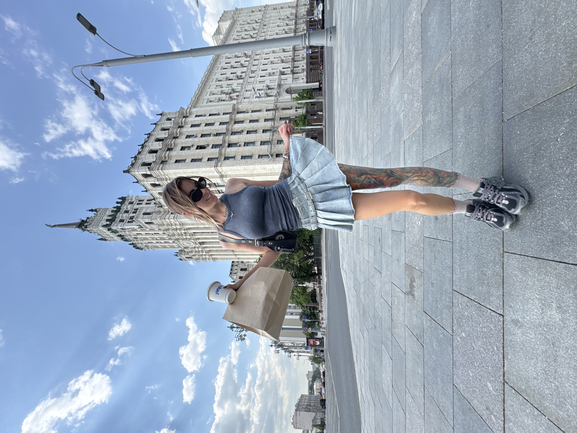 A woman stands on a gray paved sidewalk in Moscow, Russia, holding a cup and a brown paper bag. She is wearing sunglasses, a gray tank top, a denim skirt, and gray sneakers. The woman has visible tattoos on her left leg and arm. In the background, a large Stalinist skyscraper towers over the scene, set against a blue sky with scattered white clouds. A street lamp stands to the left of the building.