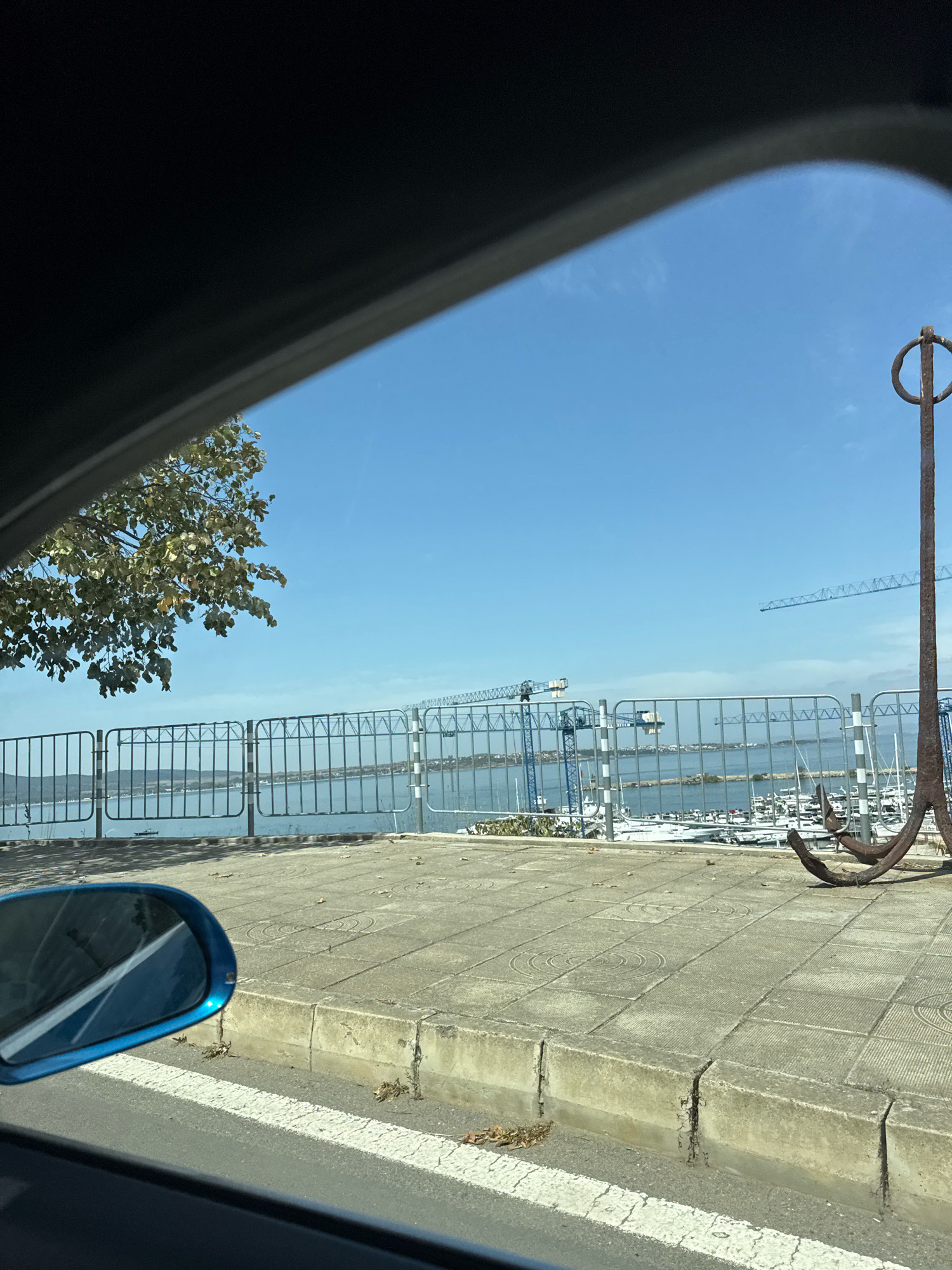 An eye-level shot captures a view through a car's side window, looking out towards a waterfront. The foreground shows a blue car side mirror and a paved walkway with square tiles. A rusted, large anchor stands prominently on the right side of the image. Beyond the walkway is a metal fence, and further out, a body of water with numerous docked sailboats. In the background, construction cranes are visible over the water, suggesting ongoing development. The sky is clear and blue. This scene is located in Sozopol, Bulgaria, specifically along the waterfront promenade.