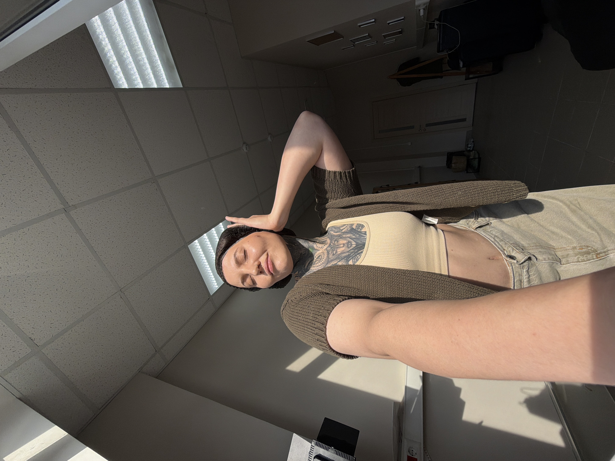 A woman with extensive tattoos covering her neck and chest is posing for a selfie in what appears to be an indoor environment, possibly an office or studio space. She is wearing a light-colored crop top, a dark knitted cardigan, and light-colored pants. Her eyes are closed, and she has her hand raised to her head. Sunlight is casting strong shadows across the room, indicating a possible window or light source. The background shows a dropped ceiling with fluorescent lighting, walls, and a doorway. The environment's architecture does not provide specific clues to pinpoint the location within the city of Chaikovsky, Russia.