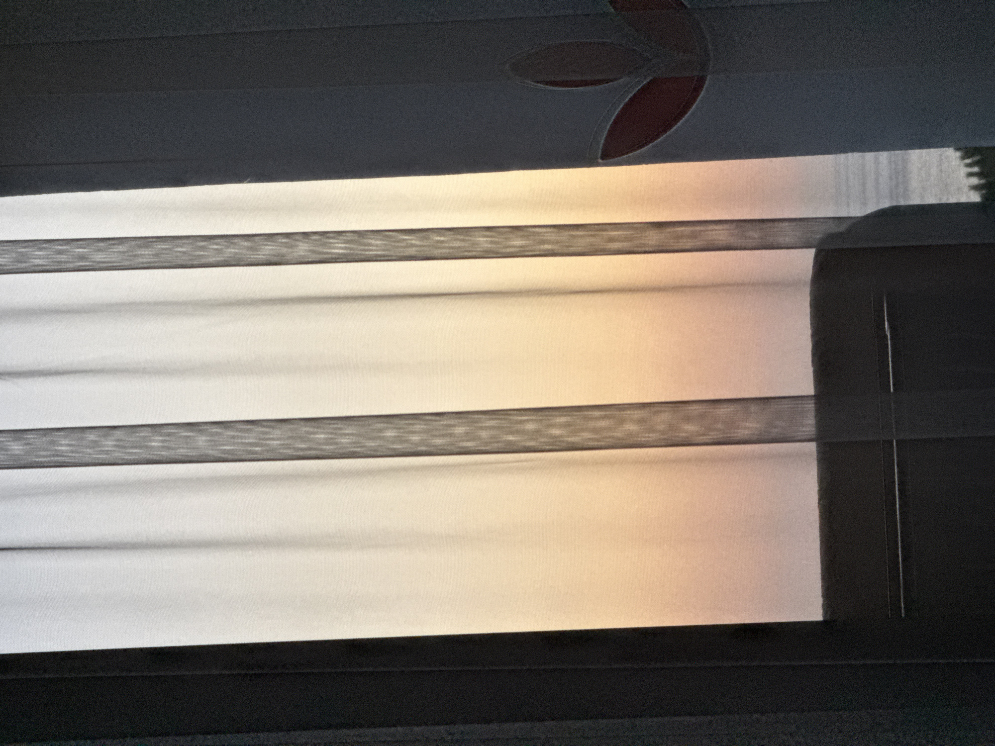 The image captures a view through a sheer curtain with vertical stripes, offering a glimpse of an ocean horizon at sunset. The curtain is illuminated by the soft, gradient light of the sky, which transitions from pale yellow at the top to a warmer orange and pink hue lower down. The dark edges of the window frame are visible on the left and right sides. On the right side, a section of a white curtain with a stylized red floral design is also present. The scene is tranquil and suggests an indoor vantage point looking out onto a coastal landscape in Menderes, Turkey, possibly from a hotel room or residence.
