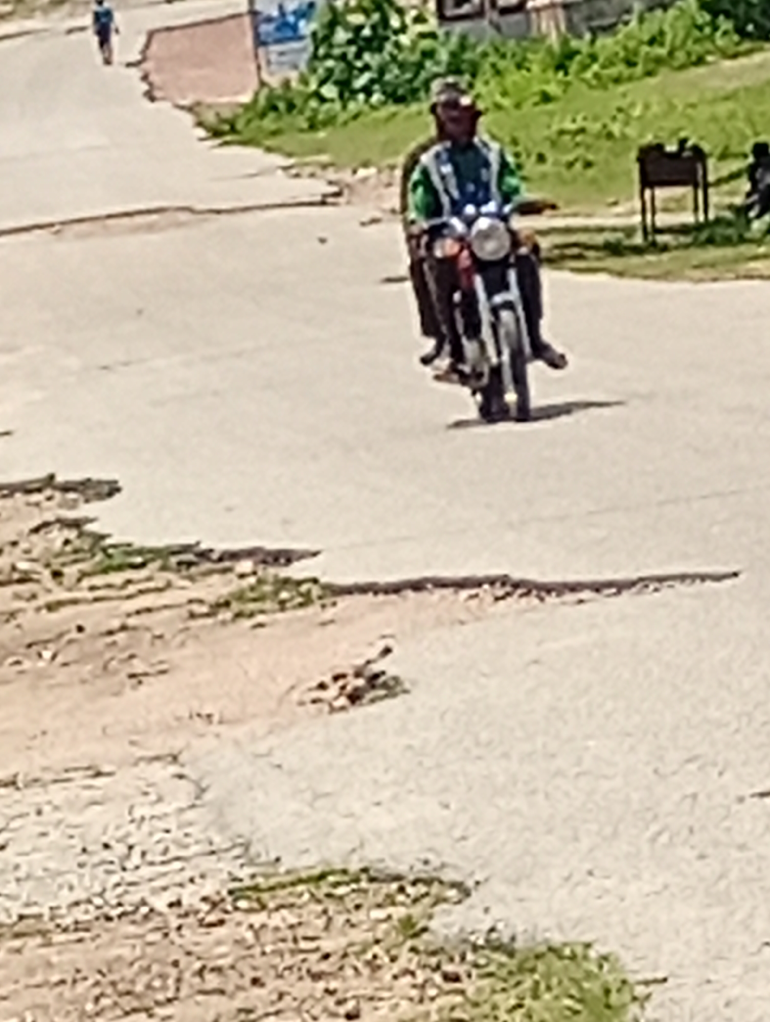 A person rides a motorcycle down a paved road in a city. The person is wearing a green and white vest and a dark helmet. The motorcycle is red and white. The road is cracked and uneven, with debris on the side. Green vegetation is visible on the side of the road, and a dark metal object, possibly a grill, is also present. In the background, a person walks away from the camera on the left side of the road. The lighting suggests it is daytime.