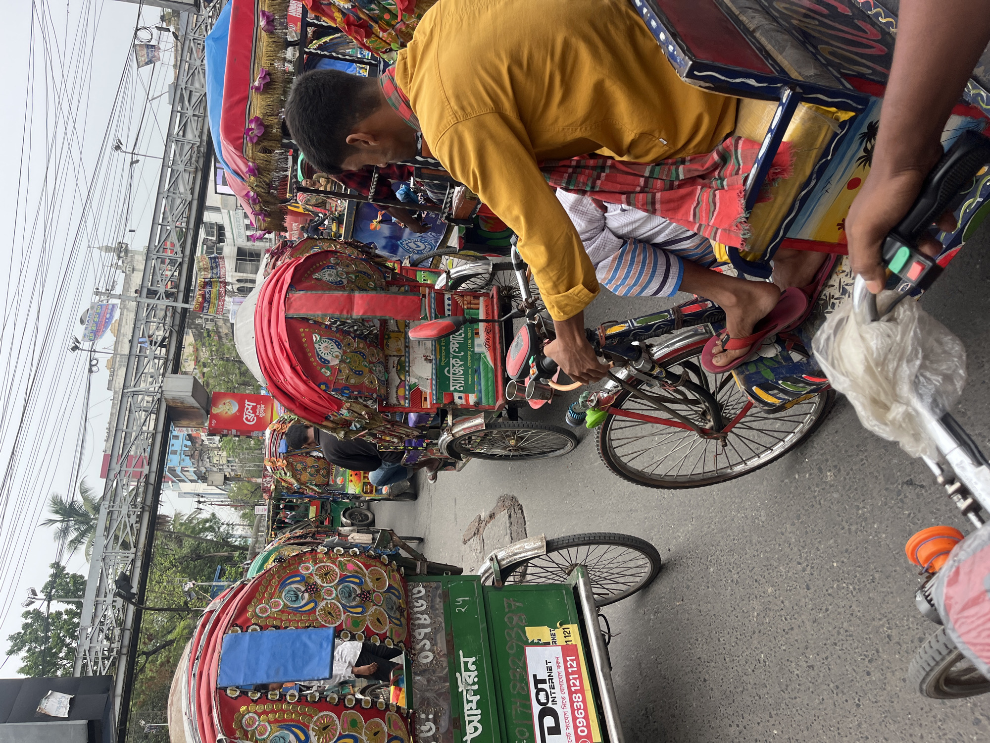 This image captures a vibrant street scene in Dhaka, Bangladesh, bustling with colorful rickshaws under an overcast sky. The perspective is from within a rickshaw, looking forward and slightly to the left.
In the foreground, a rickshaw puller wearing a yellow shirt and patterned shorts or a lungi is seen from behind, pedaling his intricately decorated rickshaw. His bare feet, clad in red flip-flops, rest on the pedals. To the right, an arm holding a rickshaw handlebar with green and red control buttons is visible, likely belonging to a passenger.
The street ahead is populated with many other traditional Bangladeshi rickshaws, each a riot of color and elaborate hand-painted designs, often featuring red, blue, and gold fringed canopies. The road surface is asphalt, showing some wear.
In the mid-ground and background, a prominent elevated metal structure, resembling an overpass or part of a metro system, stretches across the width of the image, crisscrossed by numerous overhead wires. Beyond this infrastructure, buildings, and lush green trees are visible against a bright, cloudy sky, indicating daytime.
Various texts and advertisements are visible on the rickshaws and signs:
* On a green panel of a rickshaw, words like "আফরিন" (Afrin) and "DOT INTERNET" are clear, alongside phone numbers such as "09638 121 121" and a phrase "নেট সংযোগ নিতে যোগাযোগ করুন" (Contact to get internet connection).
* Other partial texts include "ফ্রেশ" (Fresh), "ম্যাজিক স্পোে" (Magic Sport), "বিশ্বমানের ইংি" (World-class English), and "মোহাম্মদপুর" (Mohammadpur) with a phone number (01326-709471).
The image vividly portrays the dynamic street life and distinctive visual culture of urban Bangladesh.