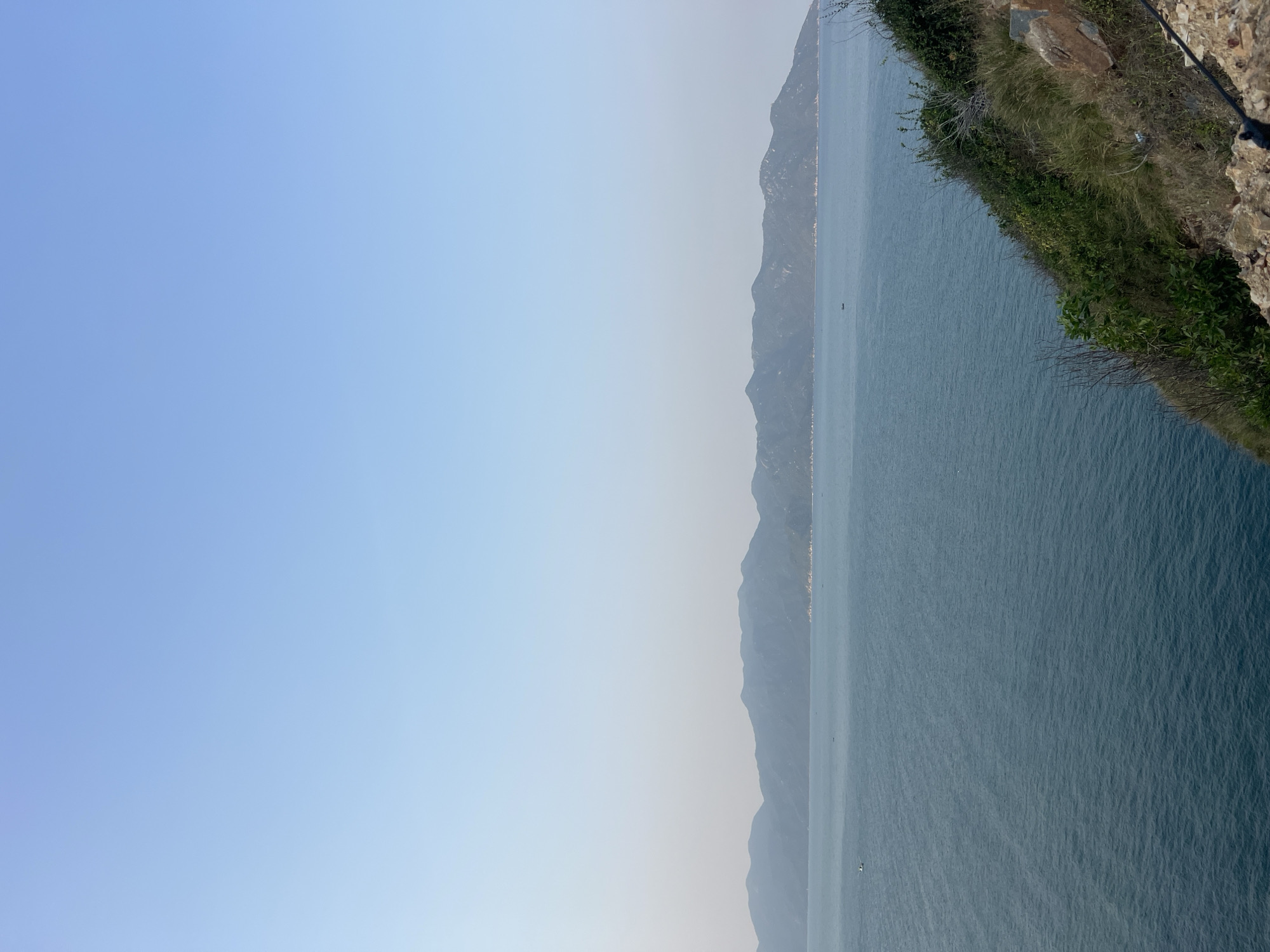 This image shows a view from a cliff overlooking the sea near Bac Natural Heritage Area Trang, Vietnam. The sky is a pale blue and takes up a large portion of the frame. A chain of mountains is visible across the water. The cliff edge is covered in vegetation. The sea is calm, with subtle wave patterns visible.