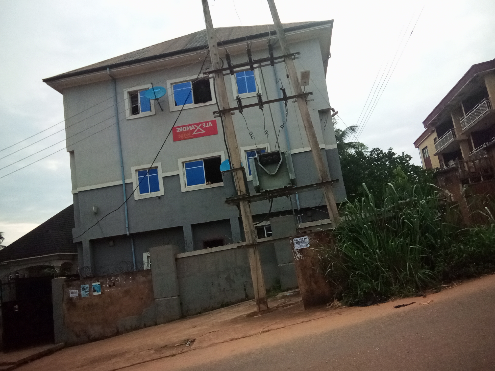 The image shows a street scene in Oko Town, Nigeria, featuring a multi-story building with a gray and white facade. The building has several windows with blue tinted glass and is adorned with a red rectangular sign that reads "ALEXANDRE" in white lettering. A satellite dish is mounted on one of the windows. In the foreground, there is a utility pole with electrical equipment, including a transformer. Adjacent to the building, there is a wall with barbed wire, and a gate with a dark metal structure is visible on the left. The ground in front of the wall is a dirt path, and the street in the foreground is paved with asphalt. Dense green vegetation grows to the right of the utility pole. In the background, another building with a balcony and railings can be seen. The sky is overcast.