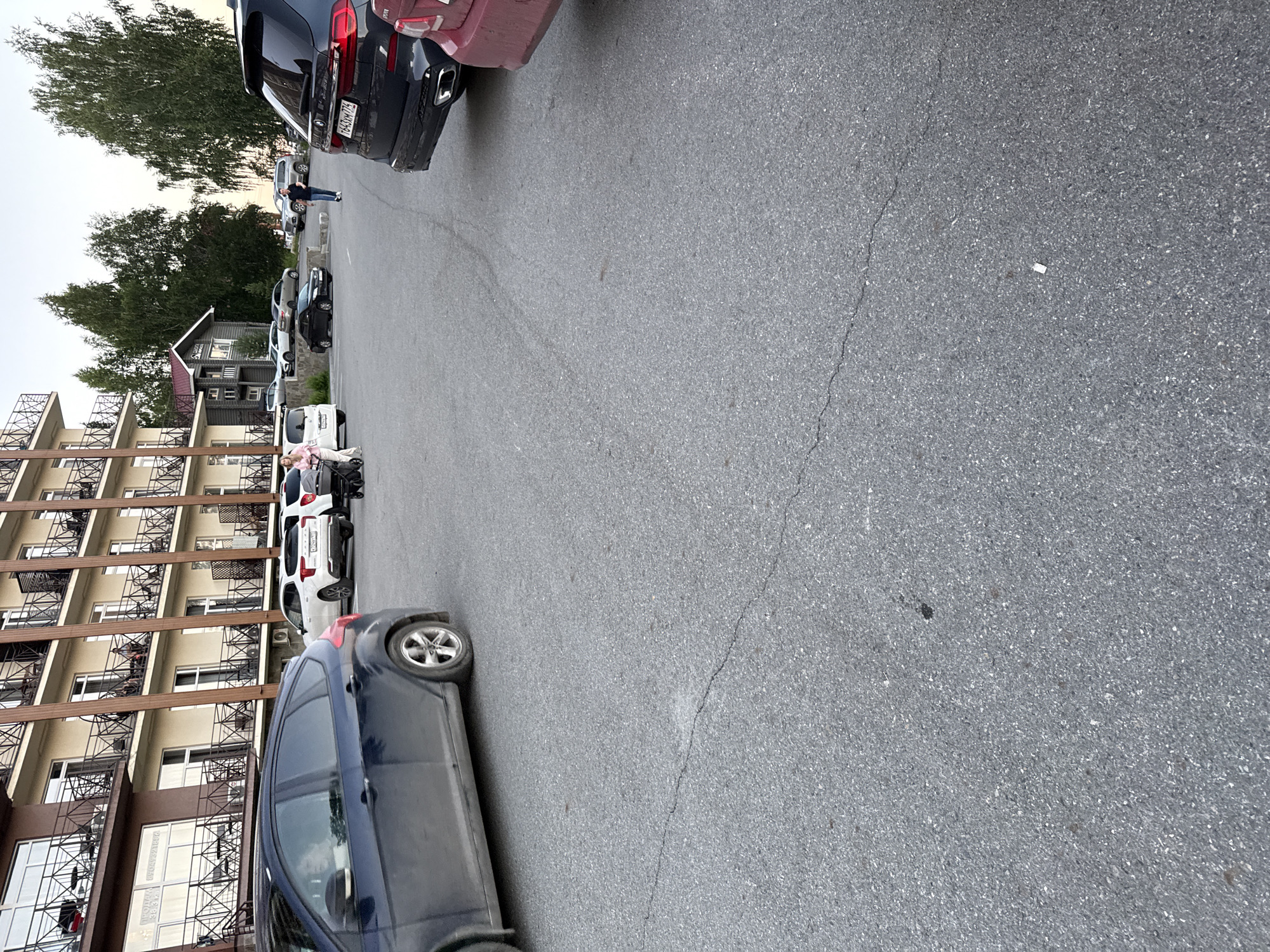The image shows a parking lot in Syrostan, Russia, in front of a multi-story building with balconies and rooms. A dark blue Ford Focus is parked in the foreground, angled towards the left. Several other cars are visible in the background, including a white Kia, a black BMW, and a red vehicle. People are also present, with one woman pushing a stroller and a man walking in the distance. The parking lot surface is asphalt with visible cracks. Trees and a wooden building are visible behind the main building. The sky is overcast.