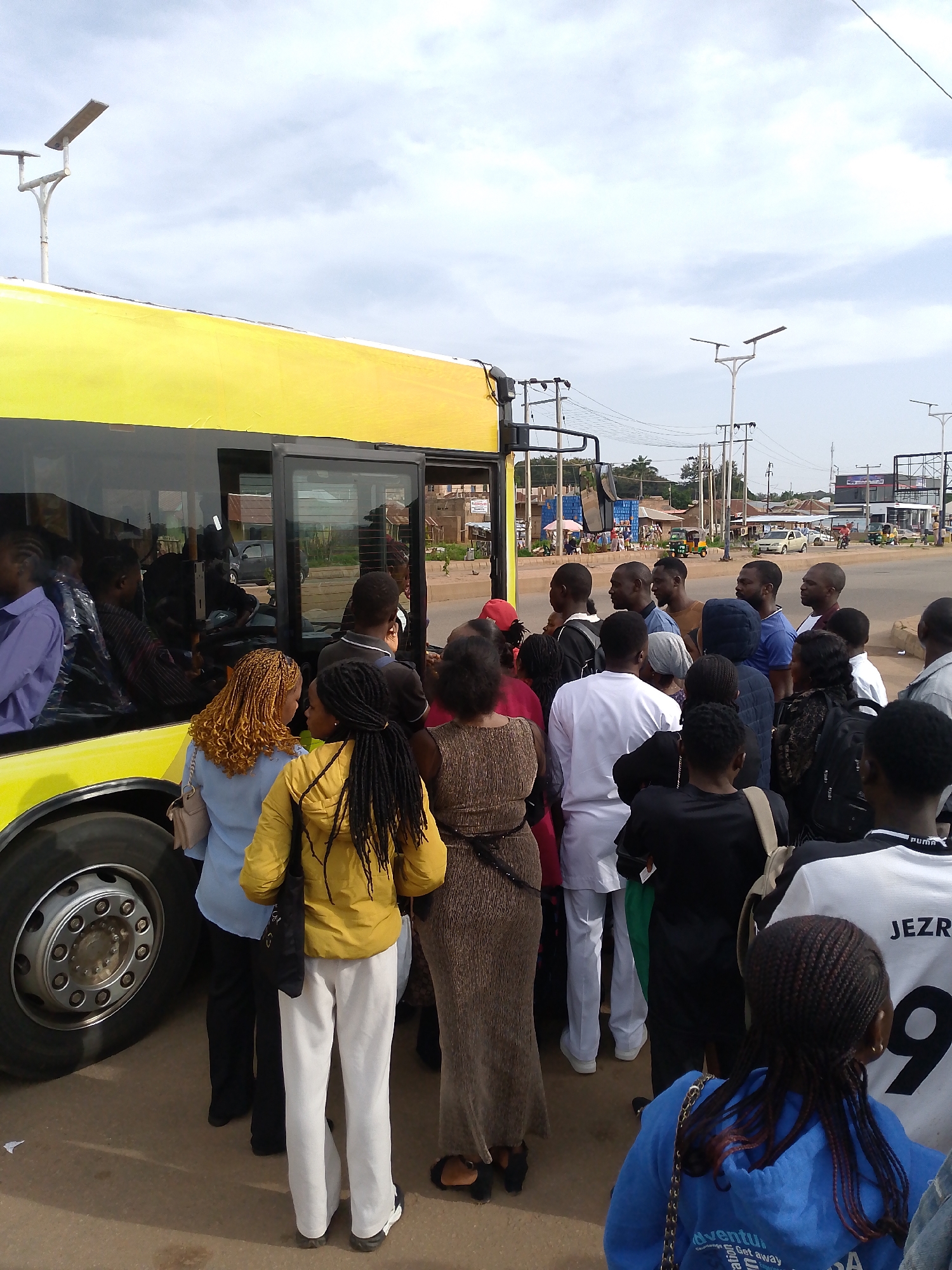 This is a wide-angle, eye-level shot of a group of people gathered around the open door of a yellow bus on a paved road. The bus has a black trim and a large, dark-tinted window. The people are diverse in age and gender, dressed in casual clothing. Some individuals are looking into the bus, others are conversing amongst themselves, and a few appear to be waiting to board or disembark.
The environment suggests a public transportation hub or a busy street corner. In the background, there are buildings, traffic lights, and utility poles, indicating an urban or semi-urban setting. The sky is overcast with scattered clouds.
The scene captures a moment of everyday life, likely during a commute or a stop for passengers. The focus is on the human interaction and the logistics of boarding or alighting the bus.
The location is Kuru Dungus, Nigeria.