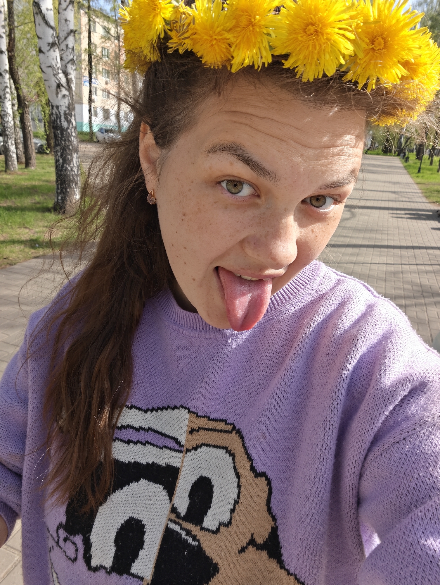 A young woman is taking a selfie outdoors. She is wearing a dandelion crown on her head and a light purple sweater with a cartoon-like design. She is sticking her tongue out. The background includes a paved path, birch trees, and a building. The location is likely in the city of Chaikovsky, Russia.