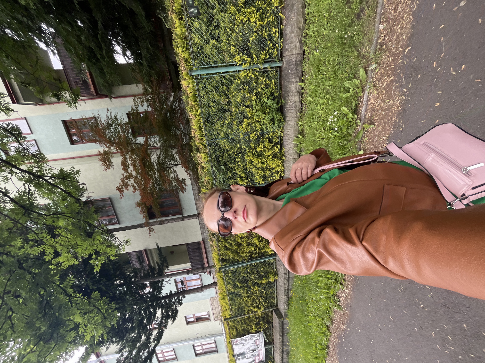 A young woman is shown in an outdoor, urban environment. She is wearing a brown leather jacket, a green top, and sunglasses. She is also carrying a pink handbag. The woman appears to be posing for the photo, looking directly at the camera with a neutral expression.
The scene is characterized by a building in the background, likely an apartment complex, with several windows and balconies visible. There is also a hedge and a fence, suggesting a somewhat manicured outdoor space. A paved walkway or road is in the foreground, with grass and scattered leaves on the side. The lighting suggests it is daytime, possibly overcast or late afternoon, as there are no strong shadows.
The image is oriented vertically, and the woman is positioned in the lower right quadrant, with her body angled away from the viewer. The overall impression is one of a casual, everyday moment captured on camera. No discernible text is visible in the image.