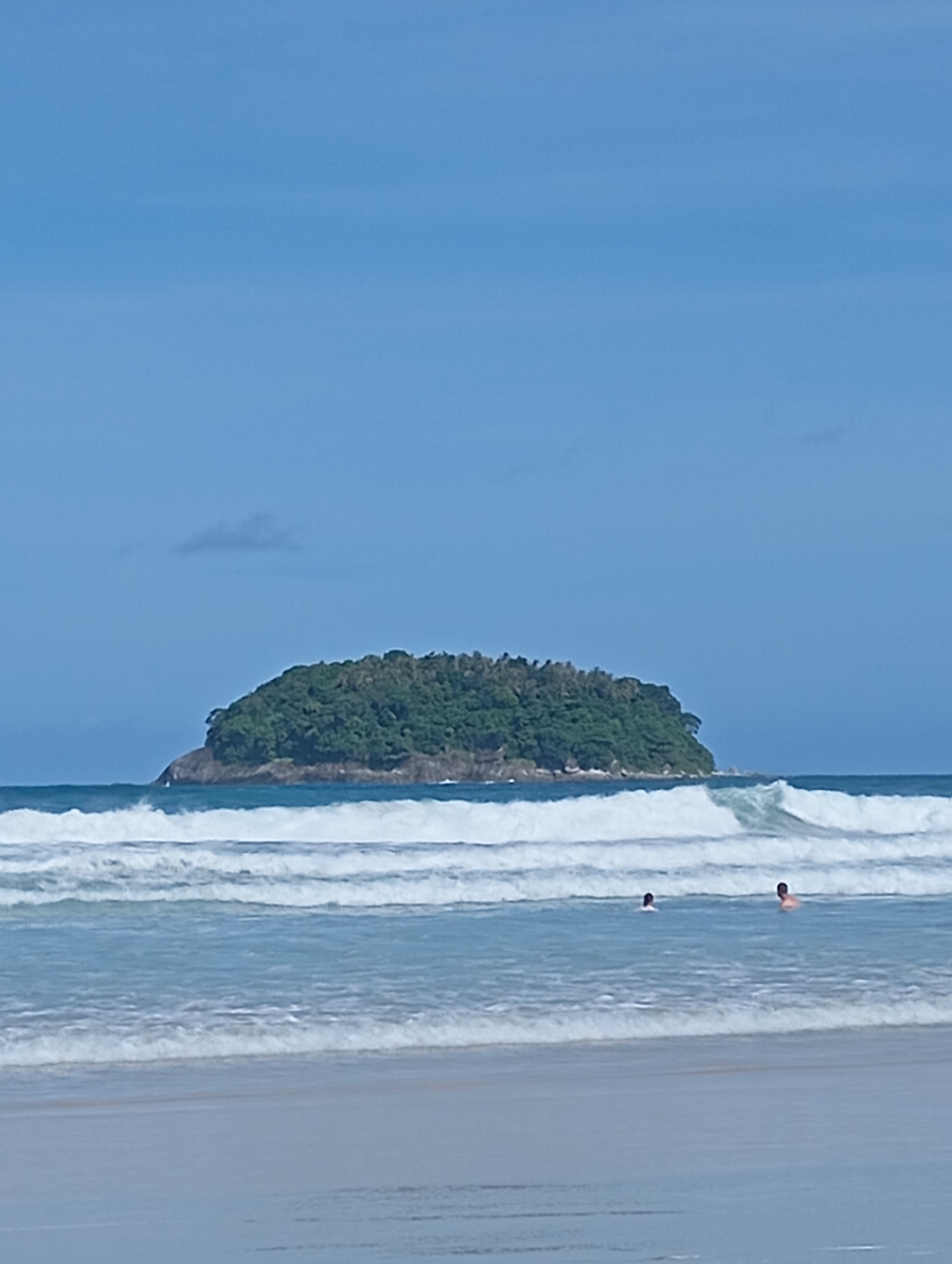 The image depicts a tropical beach scene at Karon, Thailand, with a lush, green island in the background. The ocean is a vibrant blue, with several layers of white waves rolling towards the sandy shore. Two individuals are visible in the water, swimming or wading. The sky is clear and blue, with a faint cloud formation. The overall atmosphere is serene and summery.