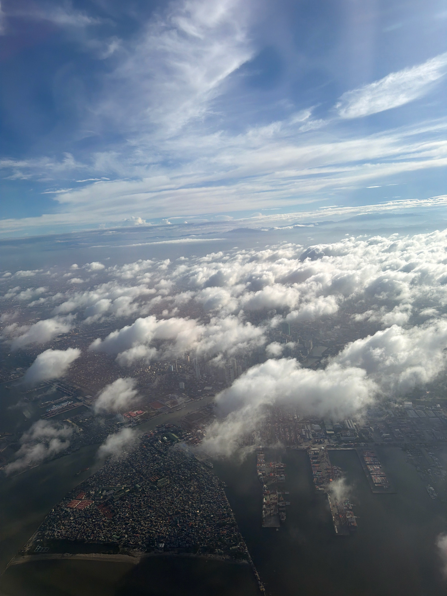 The aerial photograph captures a view of Manila, Philippines, from a high altitude. The scene is dominated by a vast expanse of clouds, with patches of sunlight breaking through to illuminate the city below.
The city itself is characterized by a dense urban landscape, with buildings clustered together. A prominent feature is the Pasig River, which winds its way through the city, reflecting the sky and clouds. Along the riverbanks and bordering the bay, a large port is visible, teeming with cargo ships and cranes. The presence of these vessels suggests a busy commercial hub.
The photograph also shows a distinct triangular landmass jutting into the water, densely populated with what appear to be residential structures. This area is a stark contrast to the more organized layout of the port and other parts of the city.
The sky is a brilliant blue, streaked with wispy white clouds, creating a dramatic contrast with the darker tones of the city. The overall impression is one of a bustling metropolis partially obscured by the natural beauty of the sky and clouds.