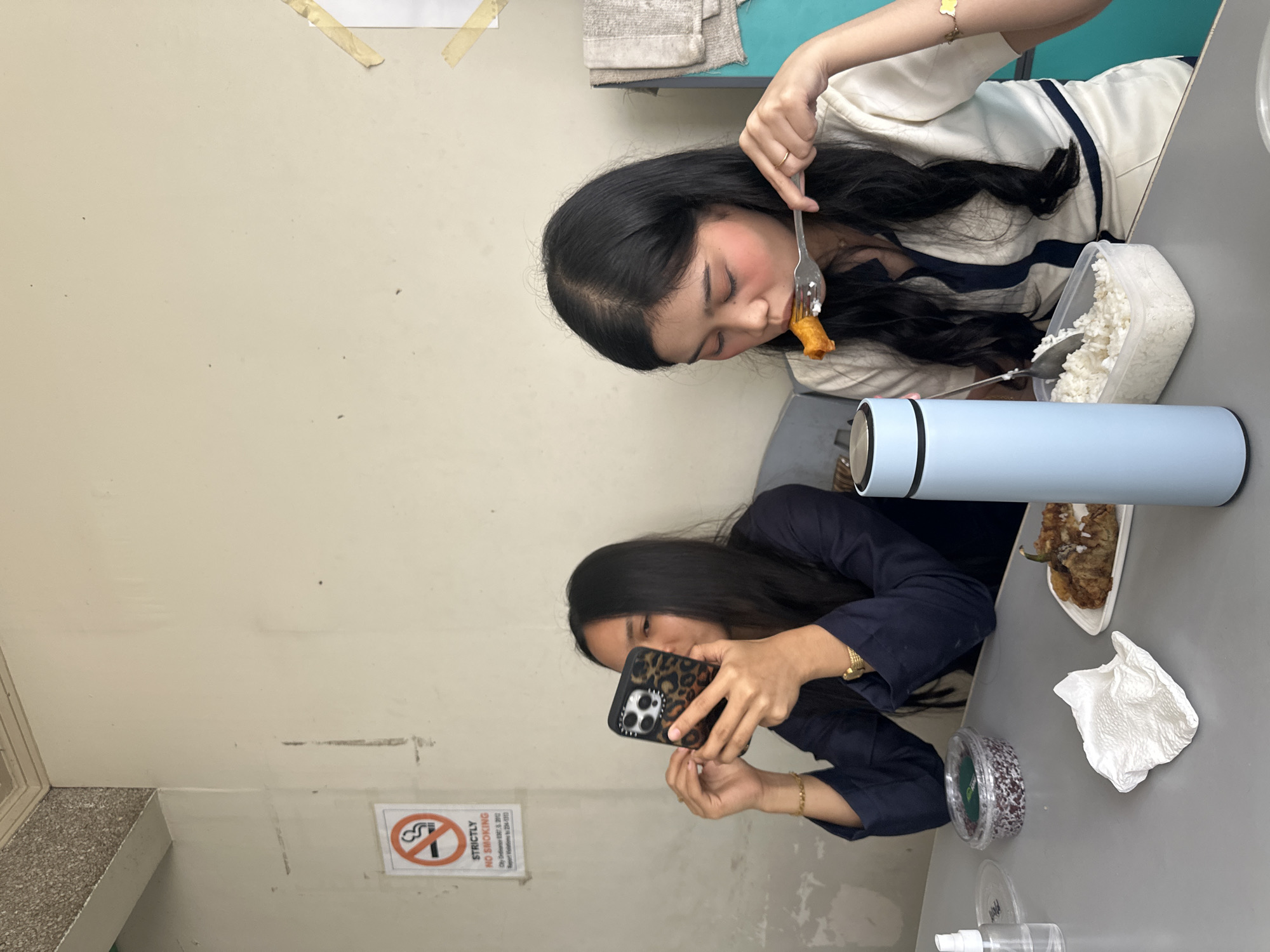 In Davao City, Philippines, two young women are seated at a gray table, engaged in dining and using their mobile devices. The woman on the left, dressed in a navy blue shirt, is holding a smartphone with a leopard-print case and appears to be looking at it. The woman on the right, wearing a white top with navy trim, is eating from a fork, bringing food to her mouth. A light blue thermos is positioned to her left, next to a clear plastic container filled with white rice. To the left of the woman in navy, a small bottle of hand sanitizer and a green-lidded container with dark contents are visible. A white paper napkin rests on the table between them, and a plate with food, possibly fried eggplant with sauce, is situated in front of the woman on the right. The background features a neutral-colored wall, a "Strictly No Smoking" sign, and part of a beige ceiling. The overall setting suggests a casual dining area within the city.