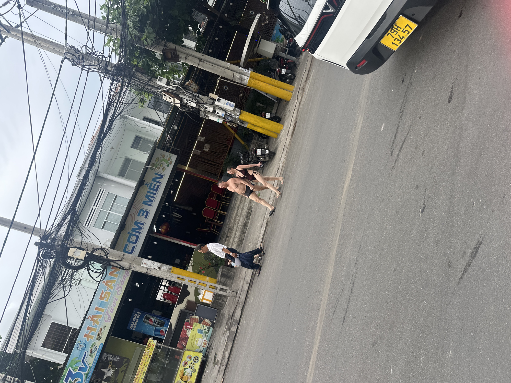 A street scene in Bac Nha Trang Ward, Vietnam shows a car with license plate "79H 134.57" parked at an angle on the right side of the image. A couple, dressed in swimwear, are walking on the sidewalk. Next to them, an older man pushes a cart. On the left, storefronts are visible with signs in Vietnamese. The electrical wires and utility poles are overhead. The image is slightly rotated clockwise.