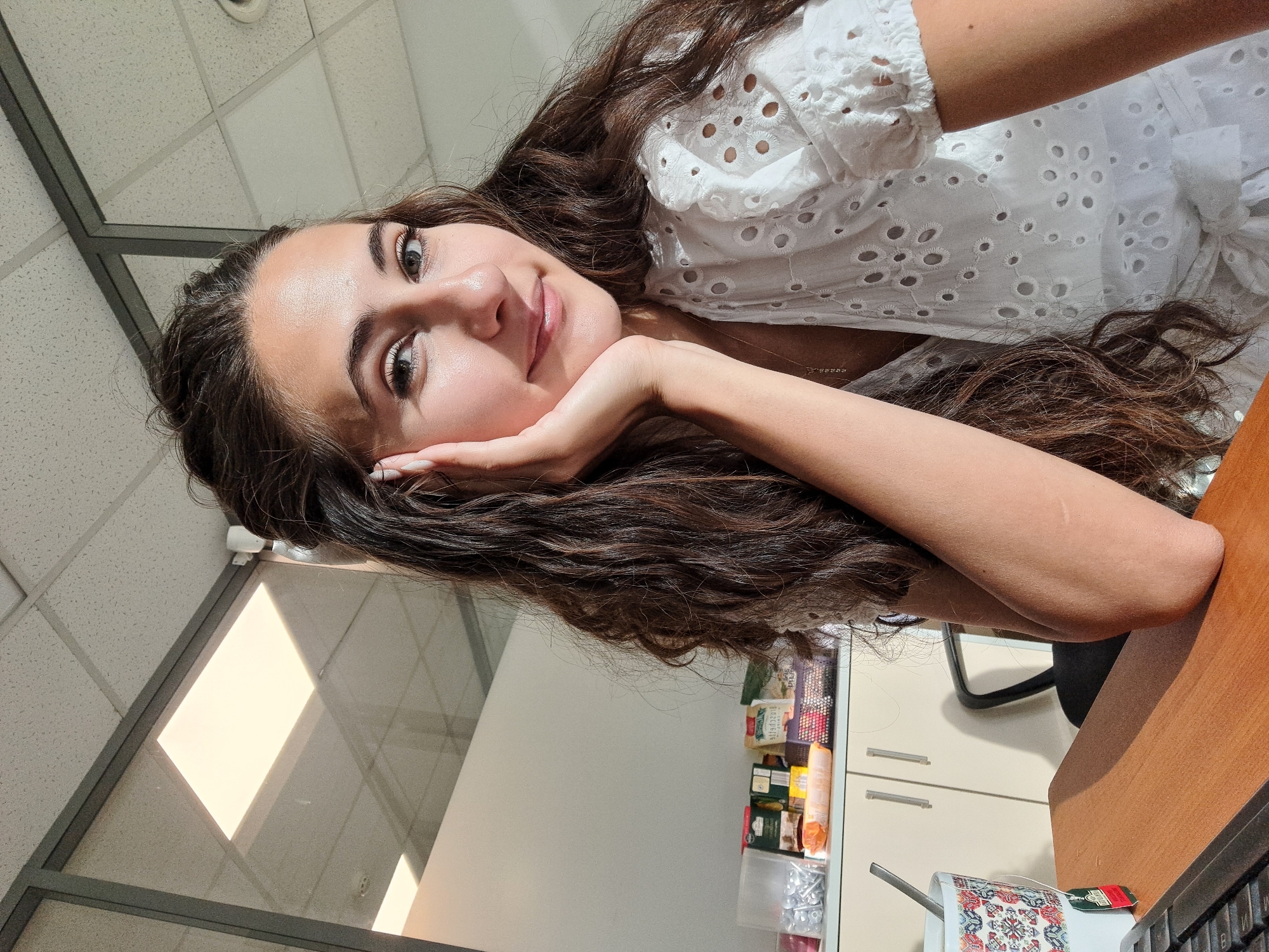 A young woman with long, wavy brown hair is seated at a desk in what appears to be an office setting, possibly in Sofia, Bulgaria. She is looking towards the camera with a slight smile, her left hand resting on her chin. The woman is wearing a white top with eyelet details. Sunlight streams in from the left, illuminating the scene and casting shadows on the ceiling. On the desk in front of her is a mug with a spoon and a tea bag tag, and part of a keyboard is visible. In the background, there are shelves with various items, including packaged goods and what looks like hardware. The environment suggests a workspace, possibly a cubicle or an office with glass partitions.
