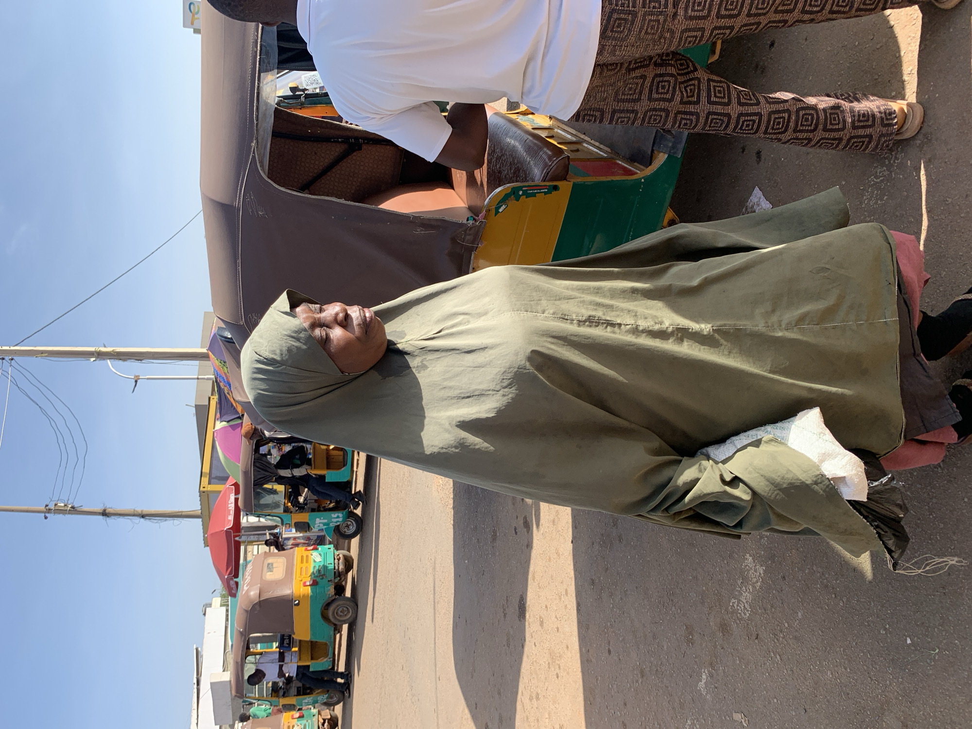 The image captures a street scene in Gura Top, Nigeria, bathed in bright sunlight, indicated by strong shadows cast on the ground. The weather is clear with a blue sky.
The central subject is a woman with dark skin, positioned in the foreground, looking to the left of the frame. She wears a full-length, sage green abaya or similar garment with a matching head covering that obscures most of her hair. Her expression is neutral, possibly pensive. In her left hand, she holds a light-colored, perhaps white, cloth and a dark plastic bag.
Behind the woman, a man in a white short-sleeved shirt is partially visible, standing next to a motorized auto-rickshaw, commonly known as a "Keke Napep" in Nigeria. The Keke Napep has a brown canvas roof and sections of green and yellow on its body. Another person's legs are visible, wearing brown trousers with a repeating square pattern and sandals, suggesting they are also standing near the auto-rickshaw.
In the background, along the roadside, several other colorful Keke Napeps are parked or moving, some featuring umbrellas that suggest market stalls or shade for vendors/passengers. Power lines run horizontally across the upper left portion of the sky. The ground appears to be a paved or unpaved road. No discernible text is visible on any objects or signs in the image. The overall scene depicts a bustling, everyday street environment.