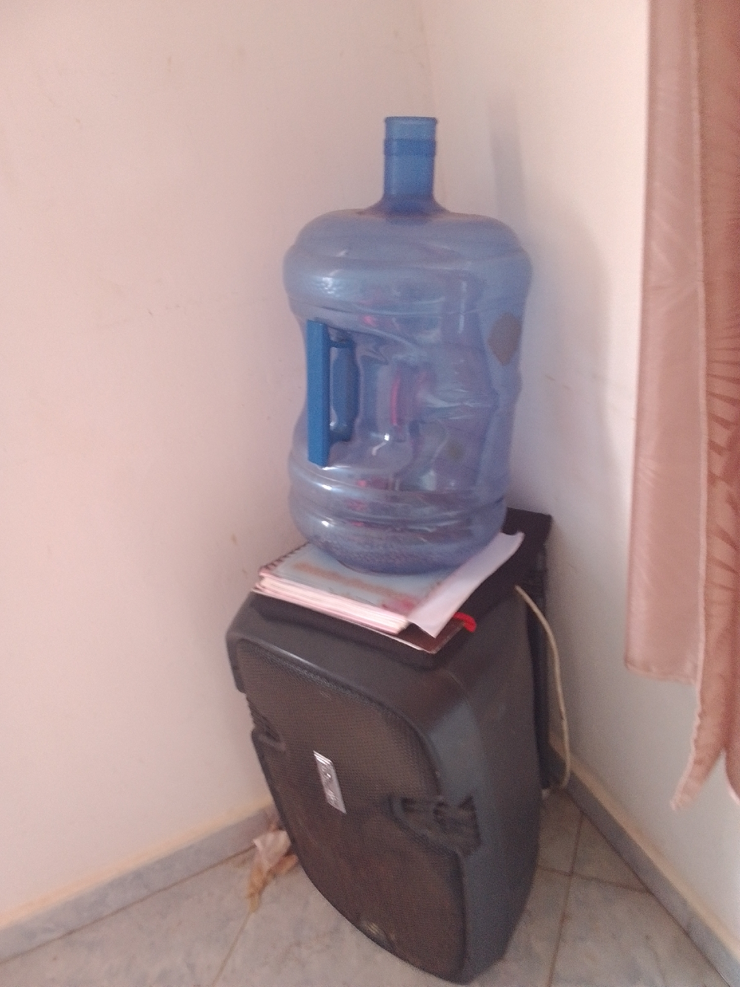 This media file captures a still life scene inside a room. The primary subjects are a large blue plastic water bottle with a handle and a black object, possibly a speaker or a storage case, positioned underneath it. The water bottle is full and appears to be a standard size for water coolers. It rests on top of a stack of papers, one of which is partially obscured by the water bottle's handle. The black object beneath has a textured surface and a metallic emblem or logo on its front. The background consists of a plain, light-colored wall and a portion of a light pinkish-brown curtain on the right side of the frame. The floor appears to be tiled. The lighting suggests an indoor setting, likely during daylight hours, though no specific time of day can be determined. There are no people visible in the frame, nor any indication of a specific activity or event. The scene is static and appears to be an organized corner of a room. No text is clearly visible in the image. The context provided indicates the location as La Tagua, Venezuela.