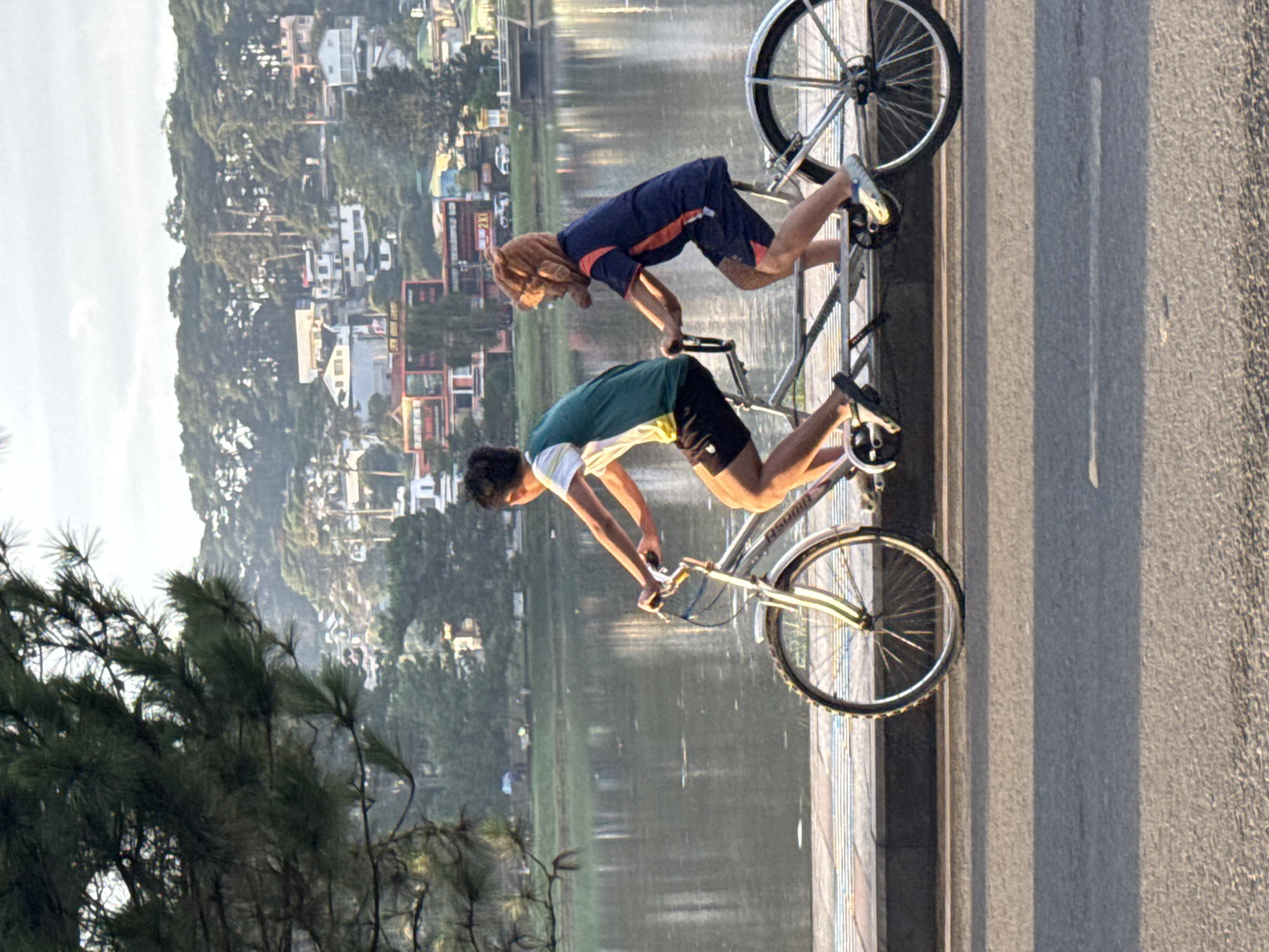 Two individuals, likely children or teenagers, are riding a tandem bicycle along a paved path beside a body of water. The person in front is wearing a teal and white t-shirt and black shorts. The person behind is wearing a navy blue shirt with orange trim and shorts, and a distinctive brown plush animal head covering. They are moving from left to right in the frame. The background shows a calm body of water with reflections of trees and buildings, and a hillside covered in dense green trees and houses. This scene is characteristic of Xuan Huong Lake in Da Lat, Vietnam, a popular spot known for its scenic beauty and recreational activities. The sun appears to be low, casting soft light and shadows.