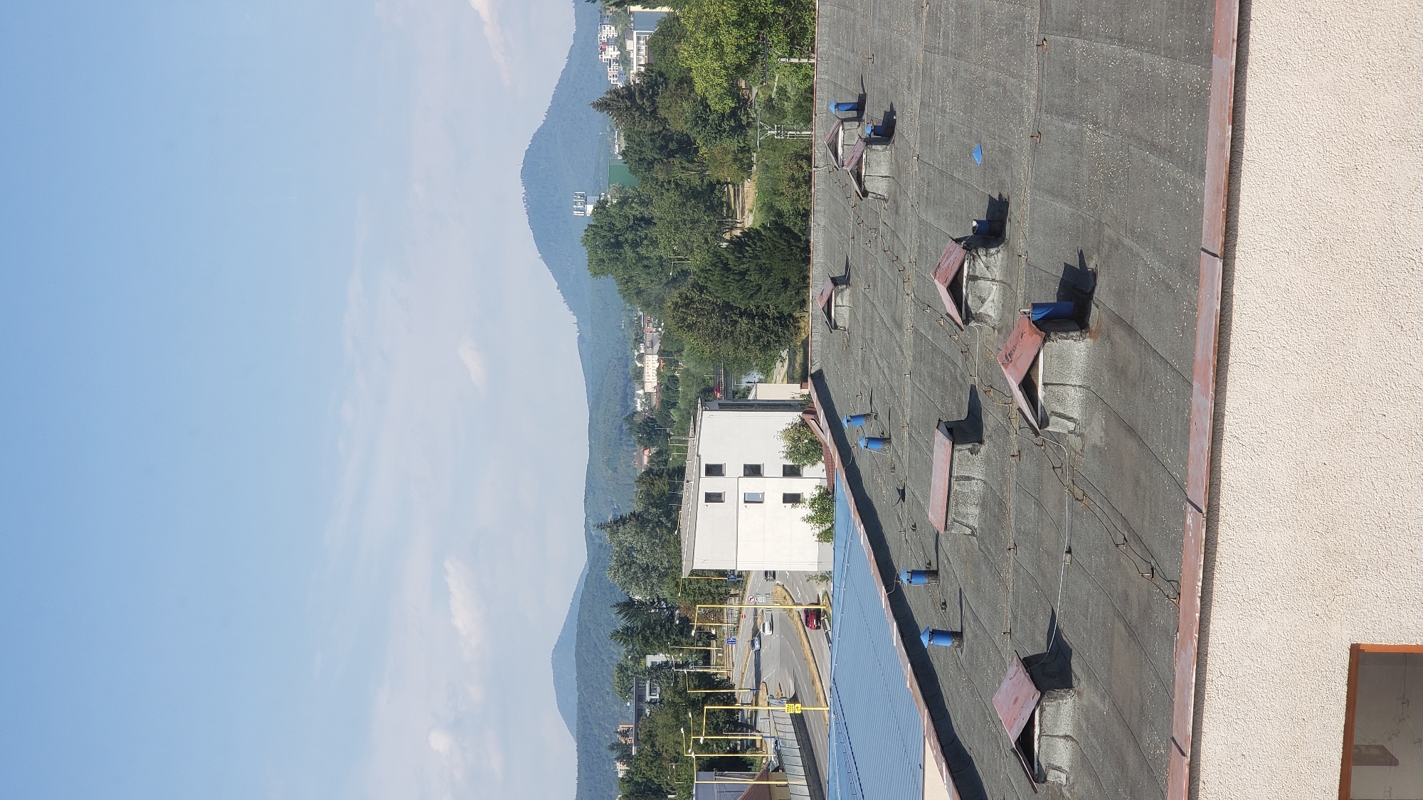 This is a daytime, eye-level, exterior shot from an elevated perspective, looking out over a city. The foreground is dominated by a flat, dark gray rooftop covered with multiple skylights, each with a reddish-brown metal frame and a peaked roof. Several blue cylindrical vents protrude from the rooftop. In the middle ground, a modern, white, multi-story building with dark-framed windows is visible. To the left of this building, a road with yellow streetlights and traffic signs runs through a residential and commercial area. Cars are driving on the road. Beyond the buildings and road, a dense line of green trees forms a backdrop, with rolling hills and mountains in the distance. The sky is a clear, light blue with a few wispy white clouds. The scene depicts a typical urban landscape with residential buildings, infrastructure, and natural elements under a clear sky, possibly in Prešov, Slovakia, given the architecture and landscape features often found there.