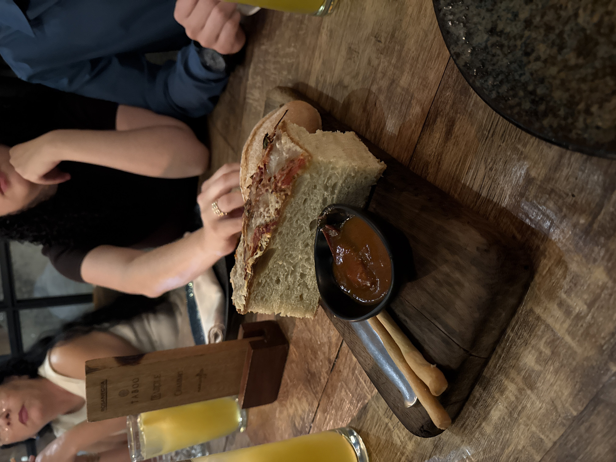 The image captures a dining scene in Mexico City, Mexico. A group is seated at a wooden table, where a wooden serving board with bread, breadsticks, and a dark-colored dipping sauce is prominently displayed. Two women and one man are visible. One woman is wearing a white shirt and a silver purse. The first woman has a gold ring and black shirt. One man is wearing a blue shirt and a watch. Two glasses with a yellow liquid, possibly juice or beer, are also visible. The restaurant's interior has a modern industrial design, as evidenced by the window frames. The overall setting suggests a casual dining experience at a restaurant in Mexico City.