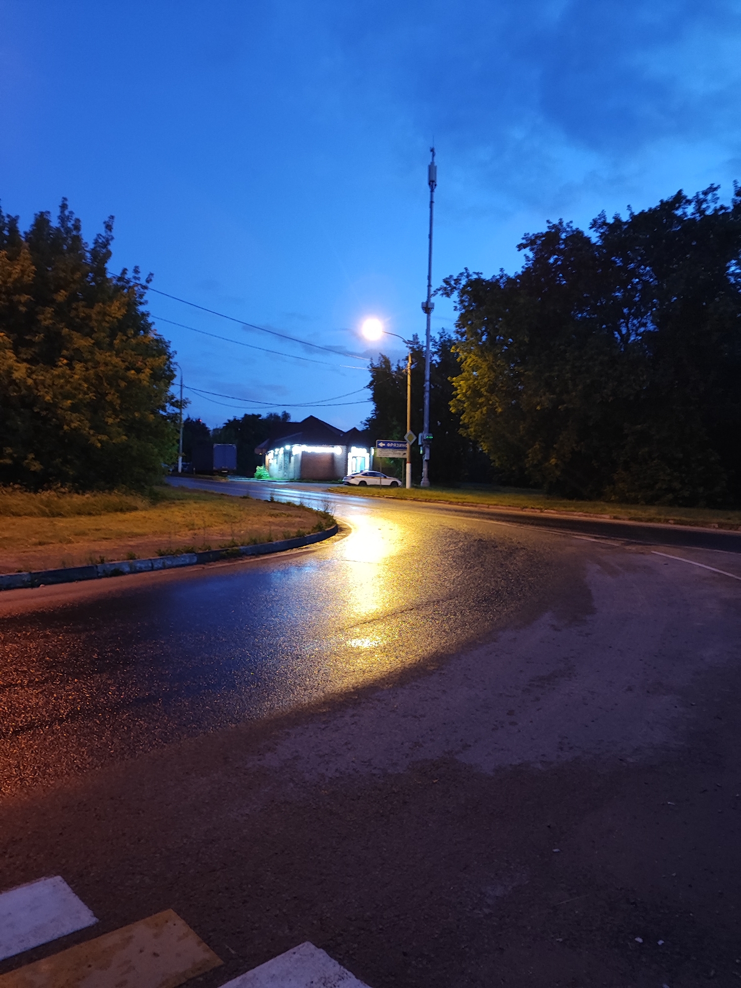 The image captures a twilight scene on a wet asphalt road in an urban or suburban environment. A curved road dominates the foreground, reflecting the light from a streetlamp and the distant lights of a building. To the left, lush green trees line a grassy embankment. In the distance, a small, illuminated building with large windows appears to be a shop or commercial establishment. A white car is parked in front of it. A tall, slender telecommunications tower stands prominently against the darkening blue sky, which is partially clouded. A streetlamp is visible on the right side of the road, illuminating a portion of the wet pavement with a bright yellow glow. Power lines crisscross the sky above the trees. The scene suggests a quiet evening after a rain, with the artificial lights creating a somewhat dramatic ambiance.