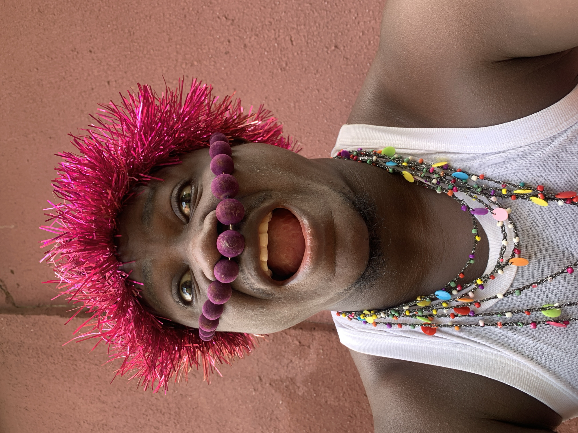 A close-up, head-and-shoulders shot shows a Black person wearing a fuchsia tinsel wreath on their head and a string of purple, fuzzy balls across their nose. The person's mouth is open, and they are wearing a white tank top with multiple layered necklaces of colorful beads. The background is a textured, reddish-brown wall. The person's skin is dark, and their eyes are wide. The image appears to be taken outdoors. The phrase "in the city of Akwanga, Nigeria" is a fictional tag and cannot be verified from the image.