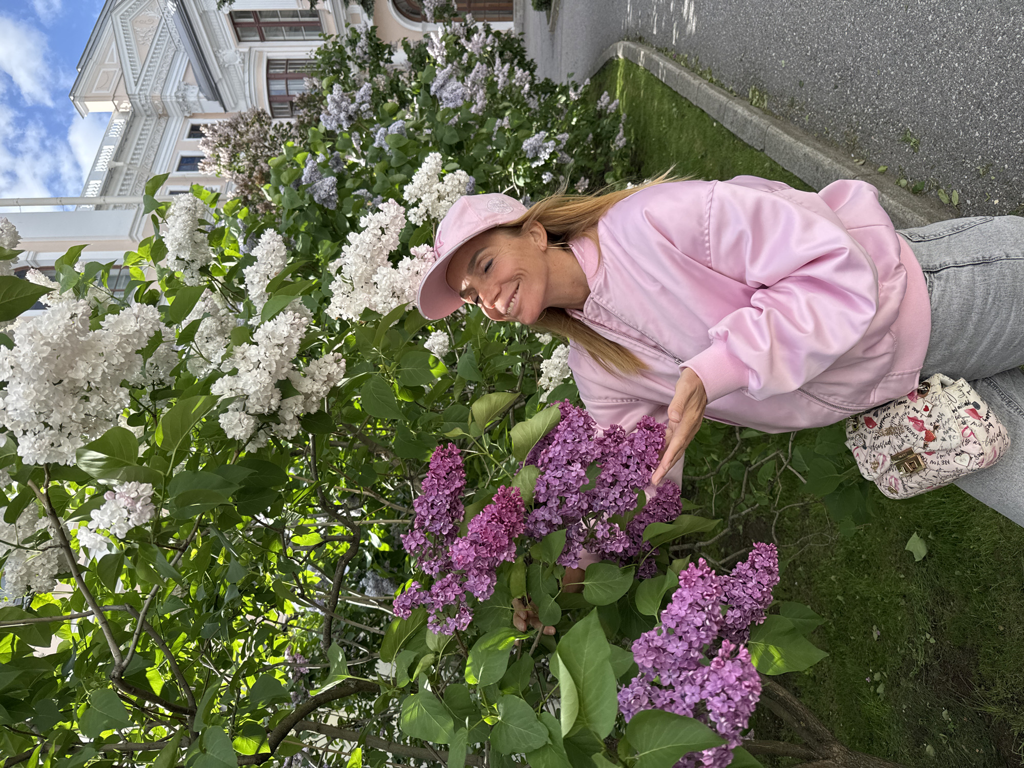 A woman in a pink bomber jacket and cap is posing next to a lilac bush. The woman is smiling and touching a lilac blossom. The lilac bush has purple and white flowers. Behind the lilac bush is a building with a light-colored facade. The scene takes place in Saint Petersburg, Russia.