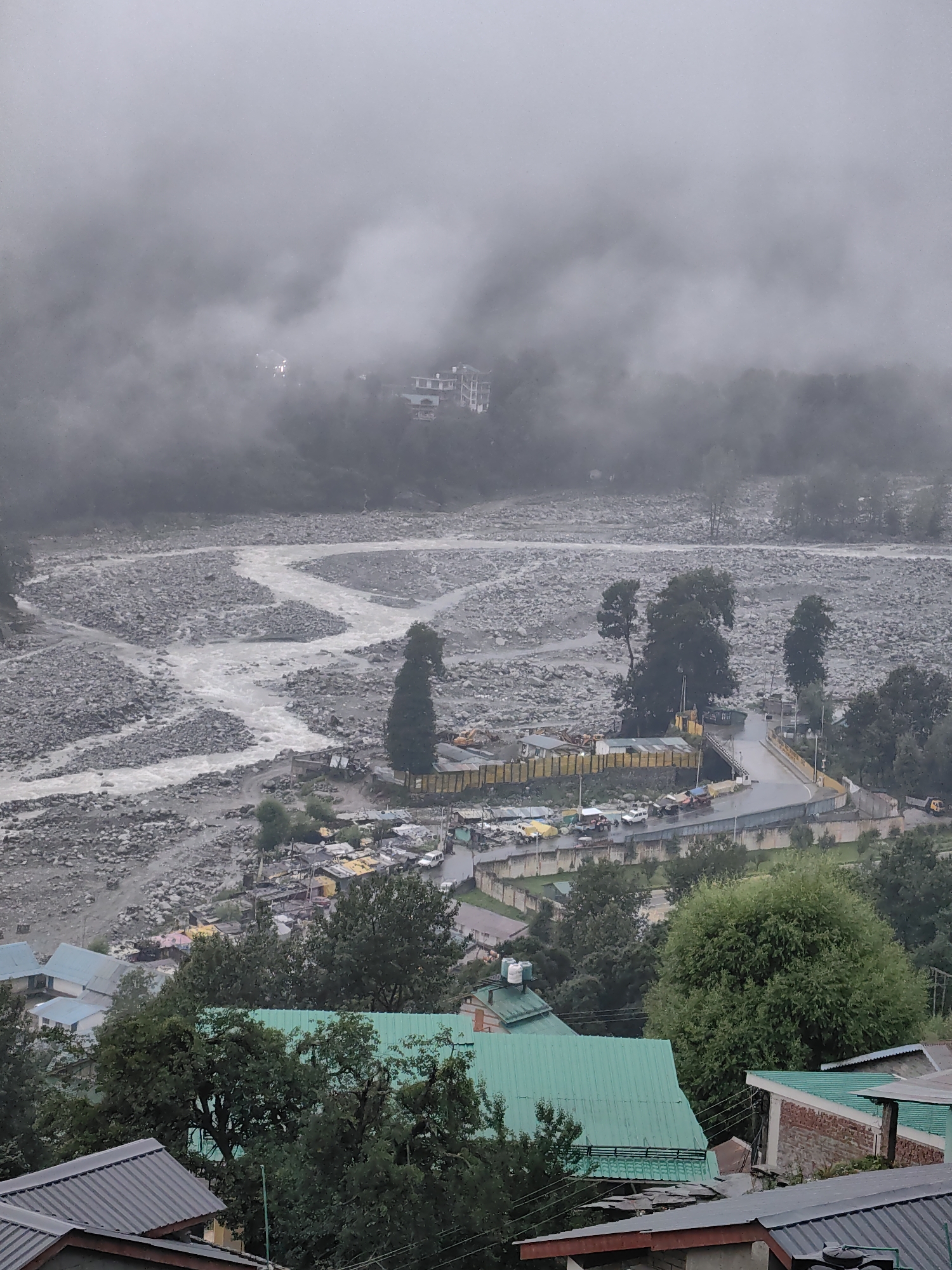 The image depicts a misty, overcast day in Vashisht, India, with a wide riverbed dominating the middle ground. The river itself is a milky white, flowing through a dense collection of grey and beige rocks. On the left bank of the river, a winding path is visible, leading towards the right side of the frame. Scattered along the riverbed and banks are small structures, possibly temporary stalls or storage units, some with yellow coverings.
A road crosses the river via a bridge, with a few vehicles visible on it, including cars and what appear to be trucks or utility vehicles. Beyond the road and bridge, a collection of buildings with colorful roofs (yellow, white, and pink) are clustered together, suggesting a settlement or market area. Several tall, dark green trees are interspersed throughout this area and the surrounding hills.
In the foreground, the rooftops of several buildings are visible, characterized by dark grey, green, and red sloped roofs. These buildings appear to be residential or commercial structures. Lush green trees partially obscure some of these foreground buildings.
The background is heavily obscured by thick, white fog or mist, which blankets the upper portion of the image and extends down into the hills, making distant structures and the landscape indistinct. Some faint lights are visible within the mist, possibly from buildings higher up on the slopes. The overall impression is one of a serene, but somewhat somber, mountainous landscape under a heavy, wet sky.