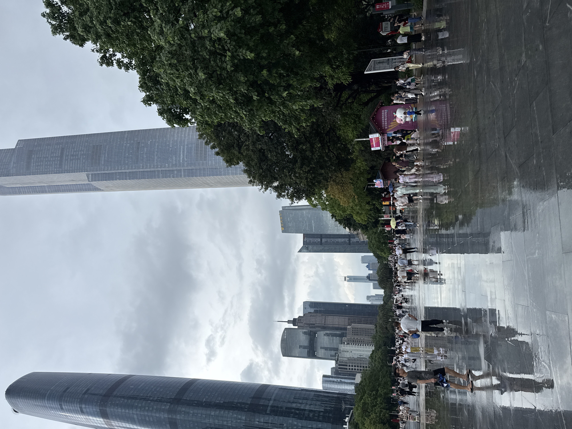 This image depicts a street scene in Tianhe District, China, with several modern skyscrapers dominating the skyline. The foreground features a wet, reflective pavement, likely from recent rain, with numerous people walking in various directions. In the middle ground, there are more buildings, trees, and a gathering of people. The sky is overcast with clouds. Specific buildings that can be identified include the Guangzhou International Finance Center and the Canton Tower in the distance. Several smaller structures and stalls are visible along the street, some with signage in Chinese. People are dressed in casual attire, and some are holding umbrellas, suggesting continued potential for rain. The overall impression is a bustling urban environment with a significant amount of glass and steel architecture.