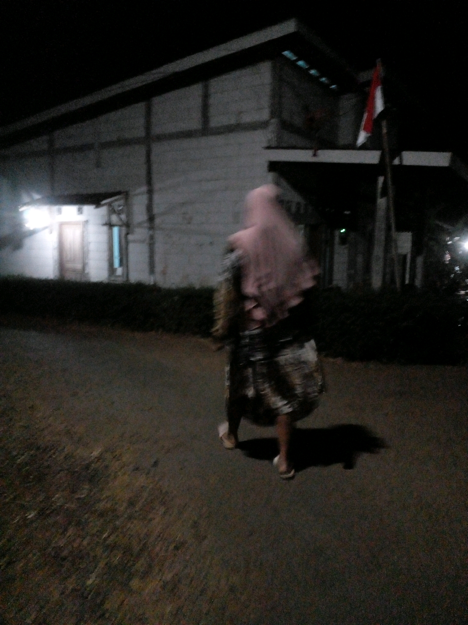 The image depicts a person walking away from the camera on an asphalt road at night. The person is wearing a light pink hijab and a patterned skirt, along with open-toed sandals. A shadow is cast behind them on the road. To the left of the person, there is a building with a lit doorway. In the background, a flag on a pole is visible, partially obscured by darkness. The environment appears to be an outdoor setting with some vegetation bordering the road. The overall lighting suggests nighttime conditions.