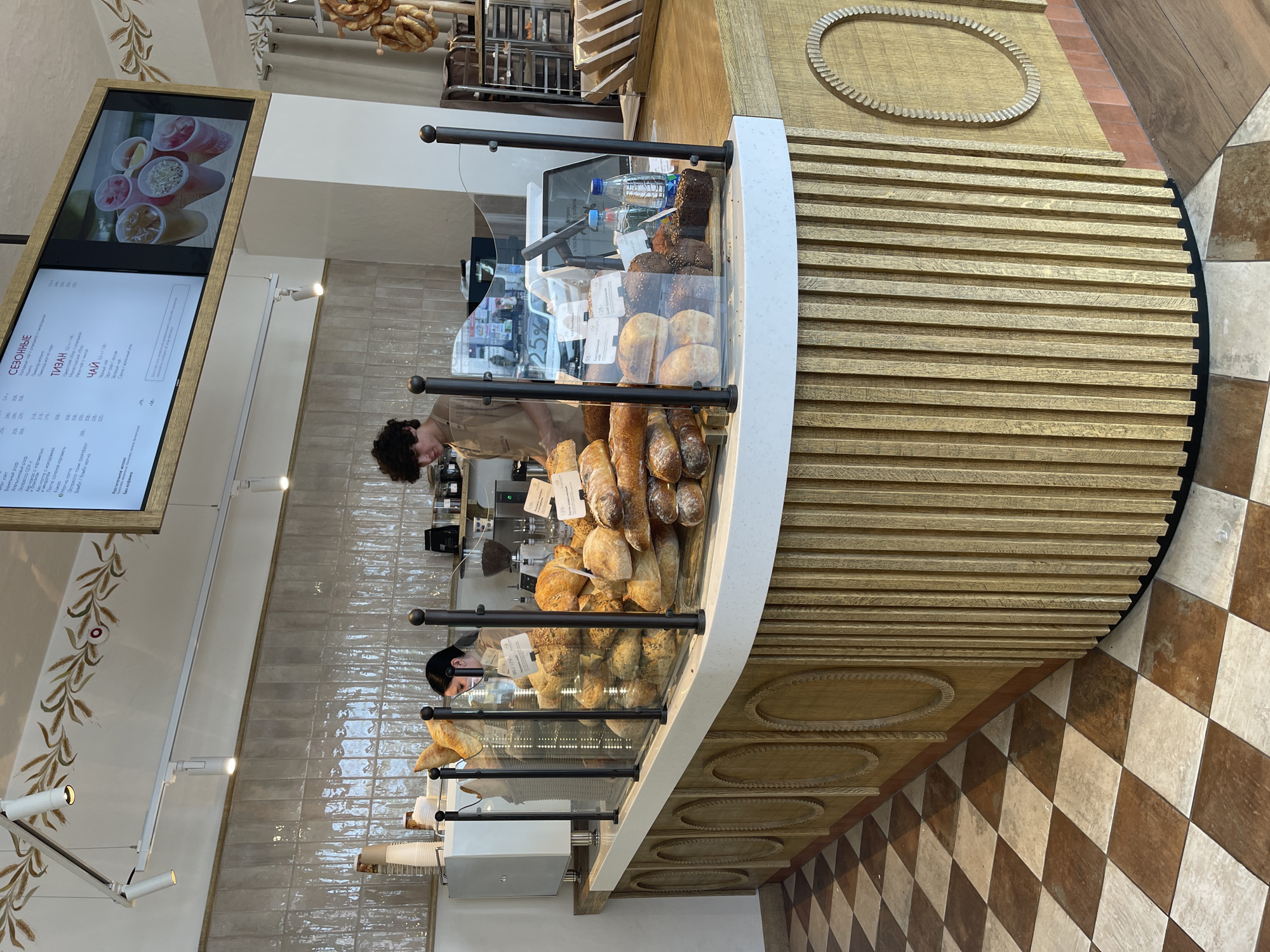A bakery counter displays a variety of fresh breads behind a glass partition. A young male employee, wearing a tan apron, stands behind the counter, partially visible as he faces away from the camera. A female employee in a tan uniform and mask is also behind the counter, looking down. The counter itself is made of wood with vertical fluting and decorative oval inlays. Above the counter, a menu board with images of drinks is displayed. The floor is tiled in a brown and beige checkered pattern. A coffee machine is visible on the left side of the counter.