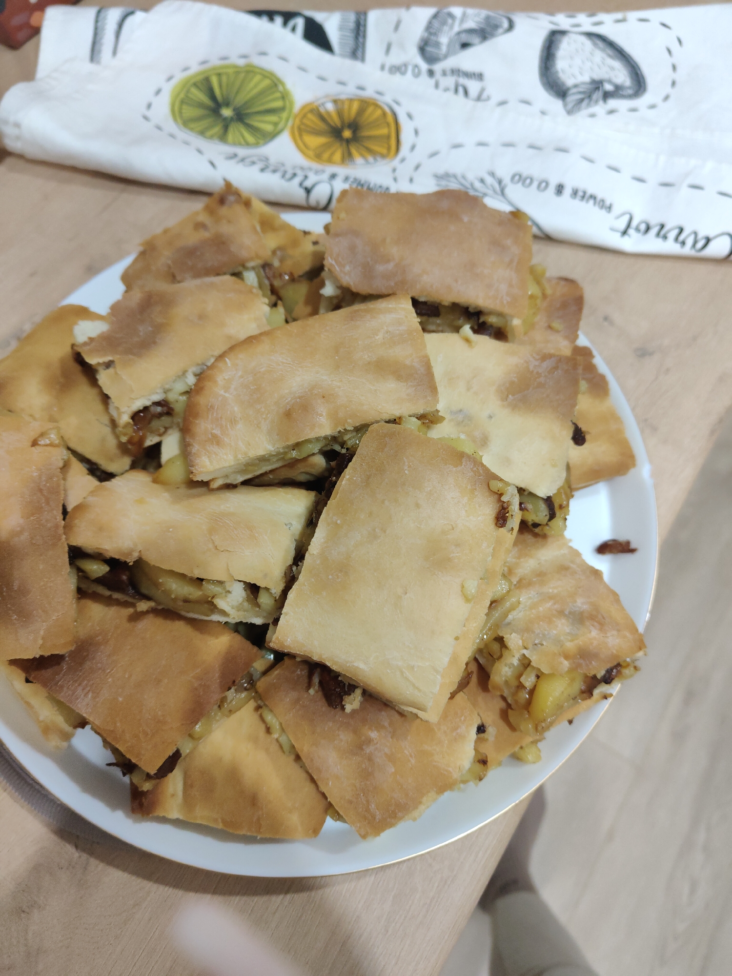 A plate of what appears to be cut pieces of pie or pastry filled with a potato and onion mixture sits on a wooden surface. The crust is golden brown, and the filling is visible in the center of each piece. A kitchen towel with illustrations of fruits and vegetables rests behind the plate. The location is Unknown, Unknown.