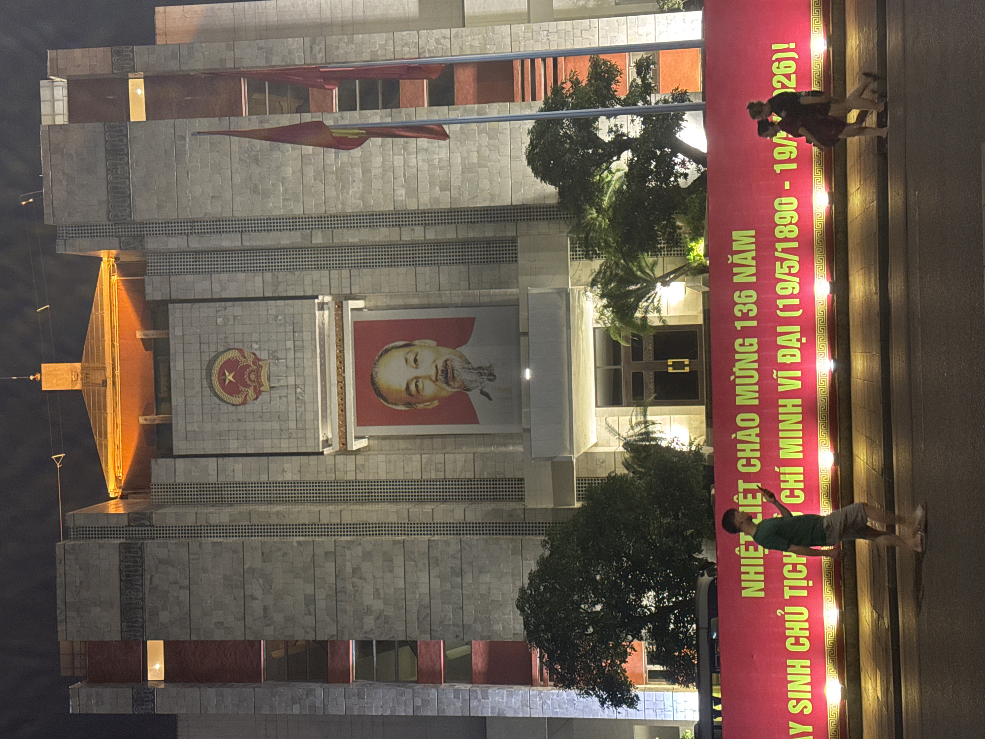 The image is of the Ho Chi Minh Mausoleum in Hà Nội, Vietnam, at night. The building is a large, multi-tiered stone structure with a large portrait of Ho Chi Minh in the center. There is a Vietnamese flag flying on the left. A large red banner is strung across the front of the building with yellow text that reads "NHIỆT LIỆT CHÀO MỪNG 136 NĂM, AY SINH CHỦ TỊCH CHÍ MINH VĨ ĐẠI (19/5/1890 - 19/26)!". In the foreground there are two people standing in the street on the right. The environment is urban and well-lit.