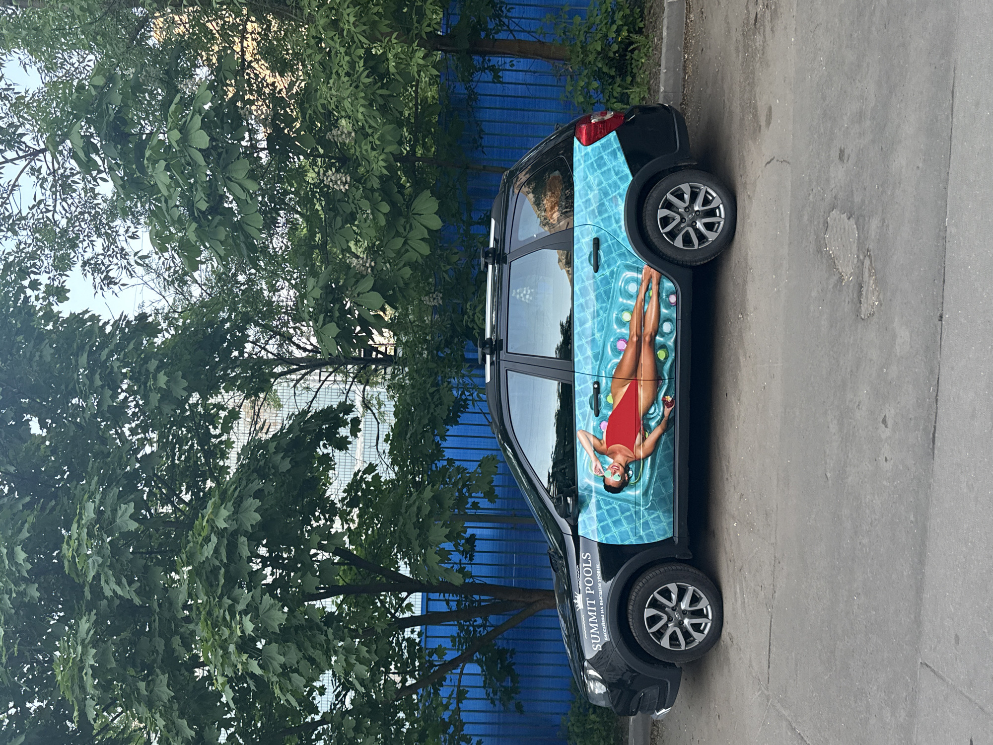 The image shows a black car parked on an asphalt road in what appears to be an urban area in Moscow, Russia. The car is partially obscured by a leafy tree on the left side. A blue fence is visible in the background. The car has a colorful advertisement for "Summit Pools" depicting a woman in a red swimsuit lounging on a pool float. The text "SUMMIT POOLS" and its translation in Russian are visible near the front of the car.