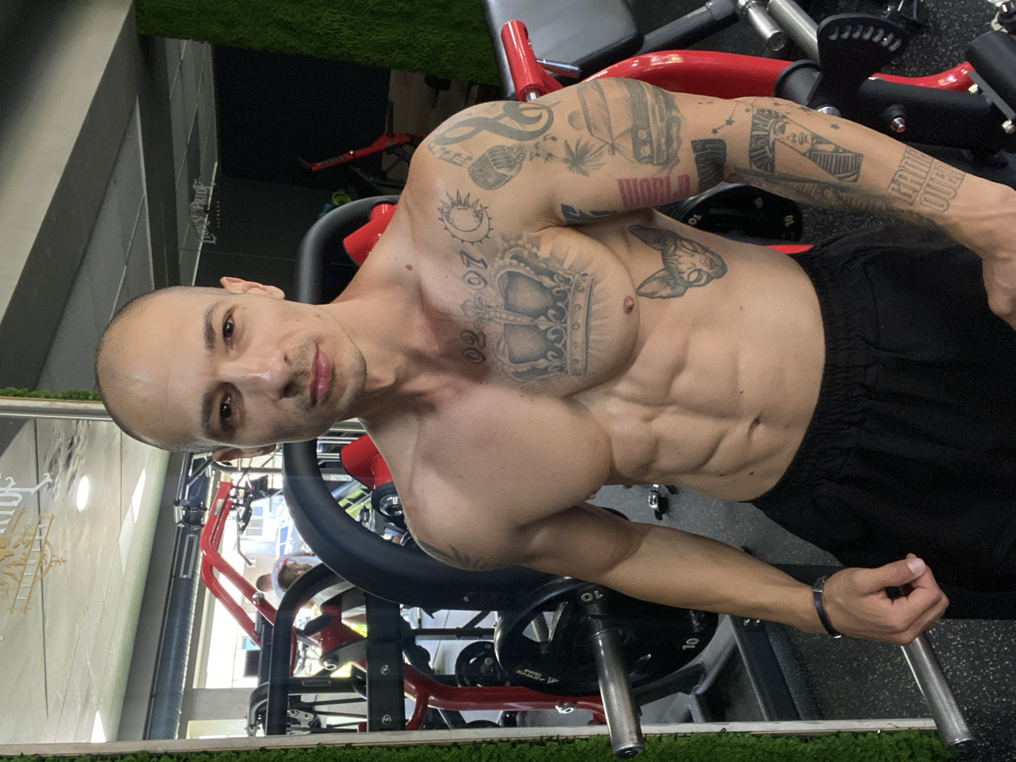 The image shows a shirtless man with numerous tattoos posing in a gym in Pleven, Bulgaria. He is bald and has a muscular physique. His tattoos include a crown on his chest, a cat's face on his abdomen, and various designs on his arms, such as a car, a microphone, musical notes, a pharaoh's head, and the phrase "GAME OVER". He is wearing black shorts and a black bracelet on his left wrist.
The gym environment is visible in the background, with weightlifting equipment, including barbells and weight plates, and a mirror reflecting parts of the gym. There are also other people partially visible in the reflection of the mirror, suggesting the gym is in use. The overall impression is a modern fitness facility.