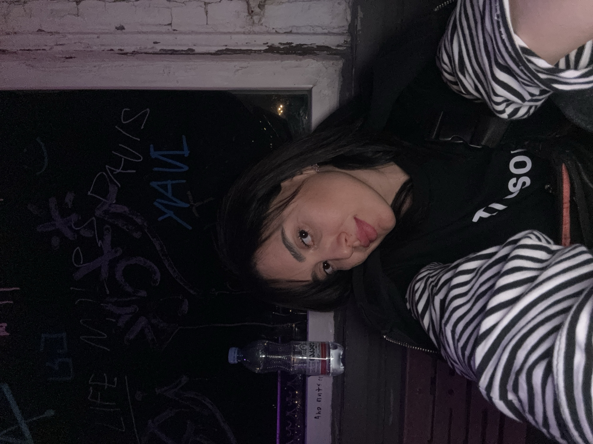 A woman is pictured in a blurry, nighttime indoor environment. She has dark hair and is wearing a black jacket with a white logo and a striped black and white sleeve. She sticks out her tongue. She stands close to an open window with graffiti on the wall that says "DAVIS," "YAVI," and "LIFE MUSIC." An unidentified bottle of water is on the window sill. There is another handwritten message on the sill, illegible but it says "Ano nuts." The lights from the city of Tbilisi, Georgia can be seen through the window, giving context to the location. The image orientation is rotated.