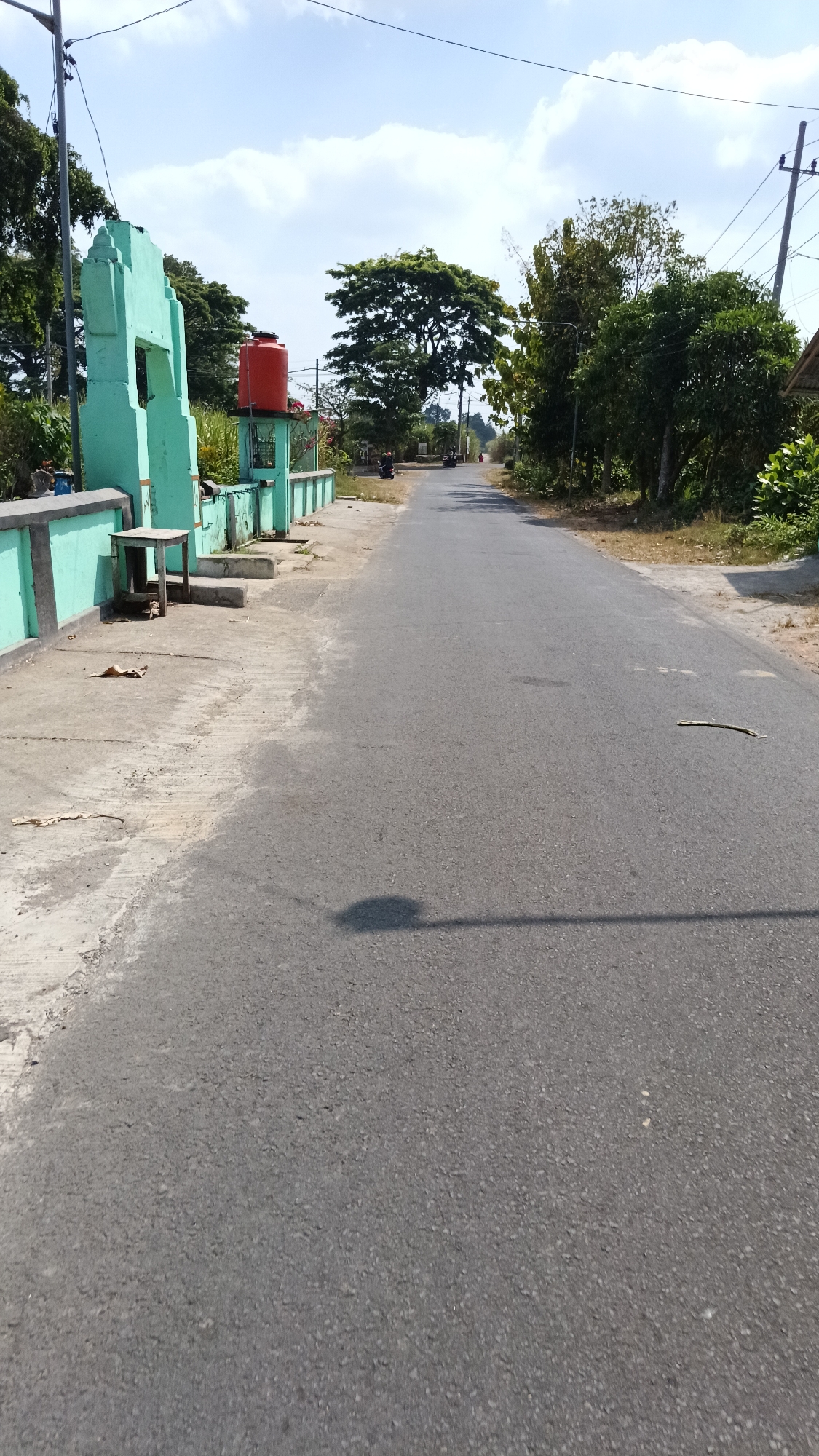 The image depicts a rural road in Wates, Indonesia, under a bright, partly cloudy sky. On the left side of the road, there is a low, light green wall with a concrete path running alongside it. Attached to the wall is a light green structure resembling a partially constructed gate or monument, with an opening in the center. To the right of this structure, a bright orange water tank is visible. A small wooden table and some concrete steps are also present near the wall. The road itself is asphalt and appears to be a two-lane street. Several motorcycles are parked or ridden on the road in the distance. Trees and vegetation line both sides of the road, with a large, mature tree dominating the middle ground on the left. The overall scene suggests a quiet, sunny day in a village environment.