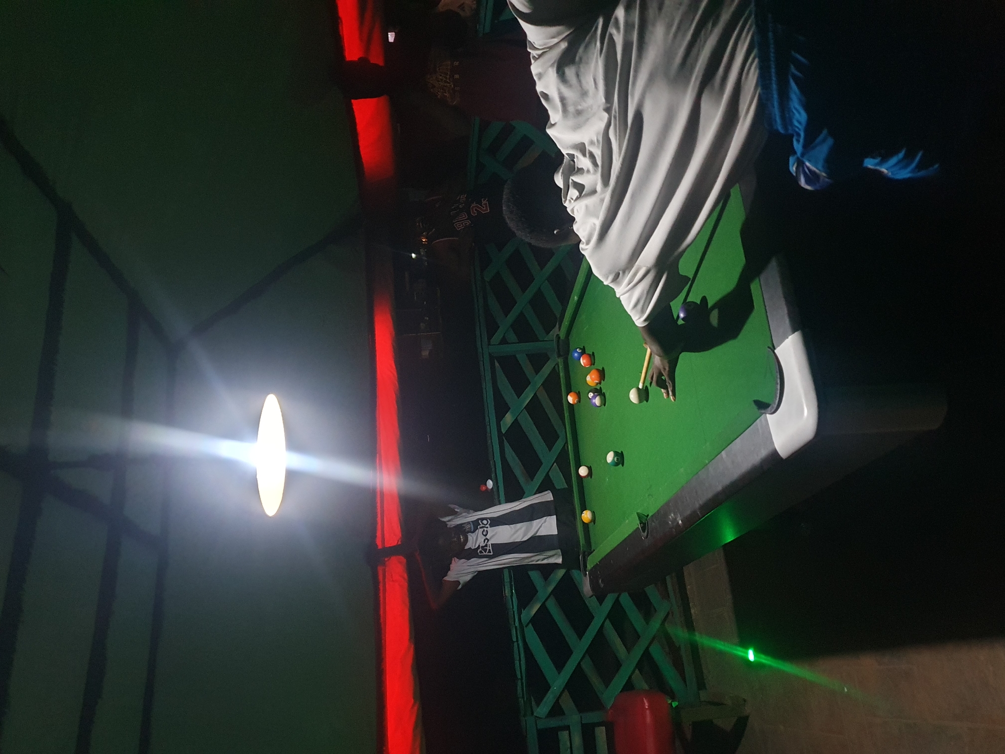 A dimly lit outdoor pool hall in Lokoja, Nigeria, features a green billiard table with several balls scattered across its surface. Two individuals are actively engaged in a game of pool. One person, wearing a white and black striped shirt, stands behind the table with their arms raised. Another person, wearing a white shirt and blue shorts, is positioned at the table, preparing to strike a ball with a cue stick. The environment is enclosed by a green wooden lattice fence and a green roof structure, illuminated by a bright overhead light and accent lighting. Red lighting is visible along the upper edge of the enclosure. A red stool is present in the lower left corner of the image, and a green laser light is visible on the floor to the left of the pool table.