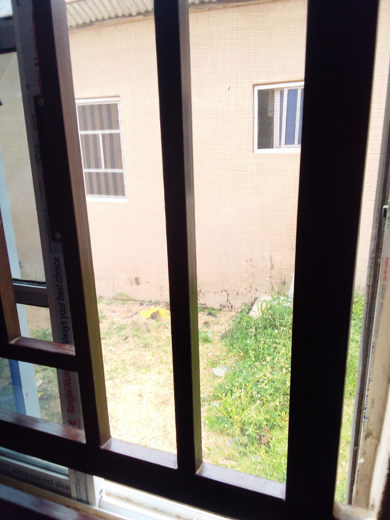 The image is taken from inside a building, looking out through a barred window. The bars are made of dark brown metal and are positioned vertically and horizontally, creating a grid pattern. Through the bars and a mesh screen, a building with a light beige facade is visible. There are two windows on this exterior wall, both with white frames. The window on the left is divided into six panes by horizontal and vertical white dividers. The window on the right is divided into four vertical panes. In front of the exterior building, there is an overgrown area with green grass and some scattered weeds. Some debris, including what appears to be a yellow plastic bag, is visible on the ground. The sky is not visible in the frame. The overall impression is of a residential or institutional building in an urban or semi-urban setting. The location is stated to be Kuson, Nigeria.