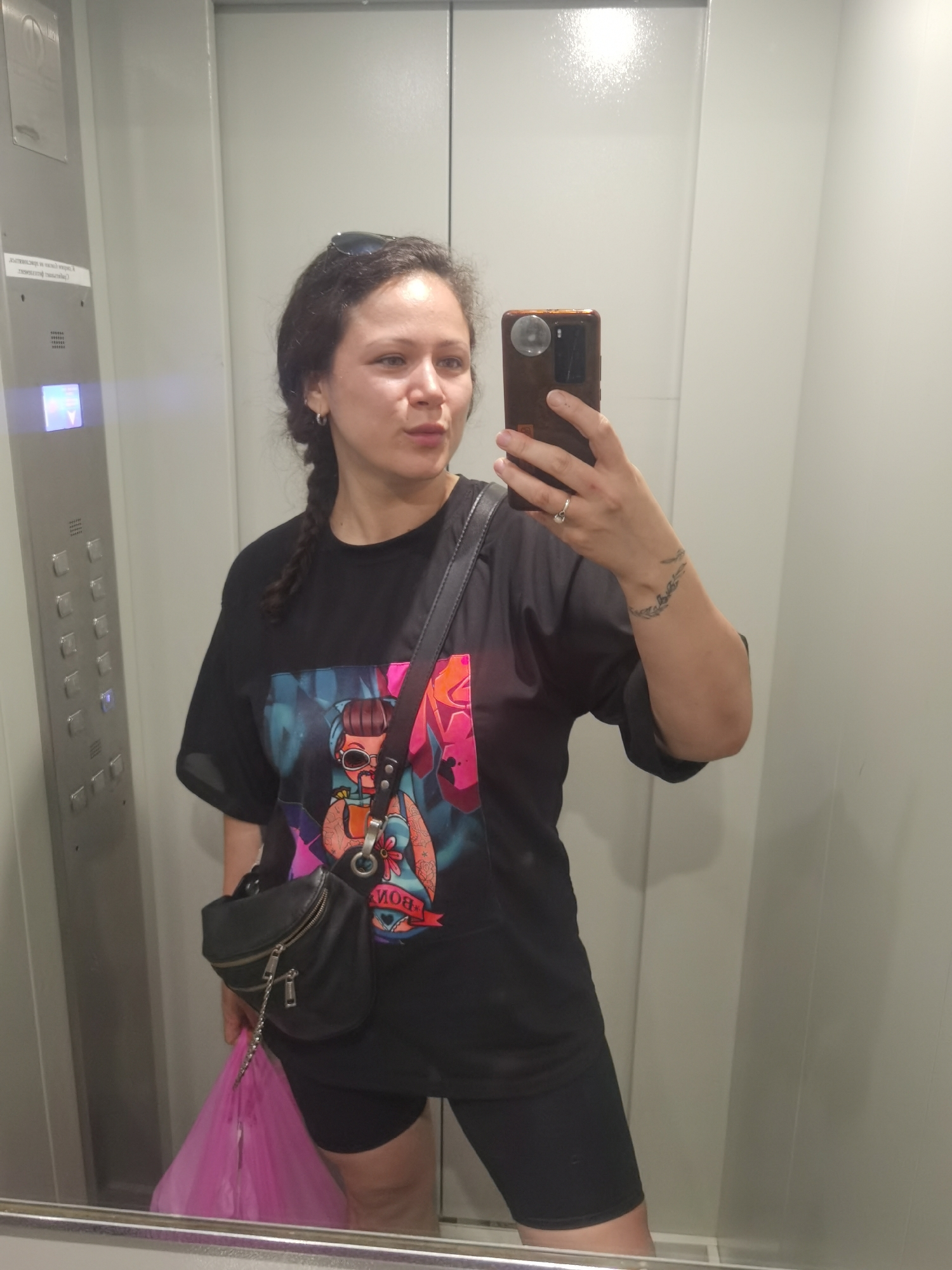 In an elevator in Chaikovsky, Russia, a young woman with dark hair braided down her back takes a selfie in a mirror. She is wearing a black t-shirt with a colorful graphic of a cartoon character, black athletic shorts, and a small black fanny pack worn across her chest. She has sunglasses on her head and a silver ring on her left hand. A bright pink plastic bag is visible hanging from her left hand. The elevator walls are light gray, and an elevator control panel with buttons and a display screen is visible on the left side of the image.