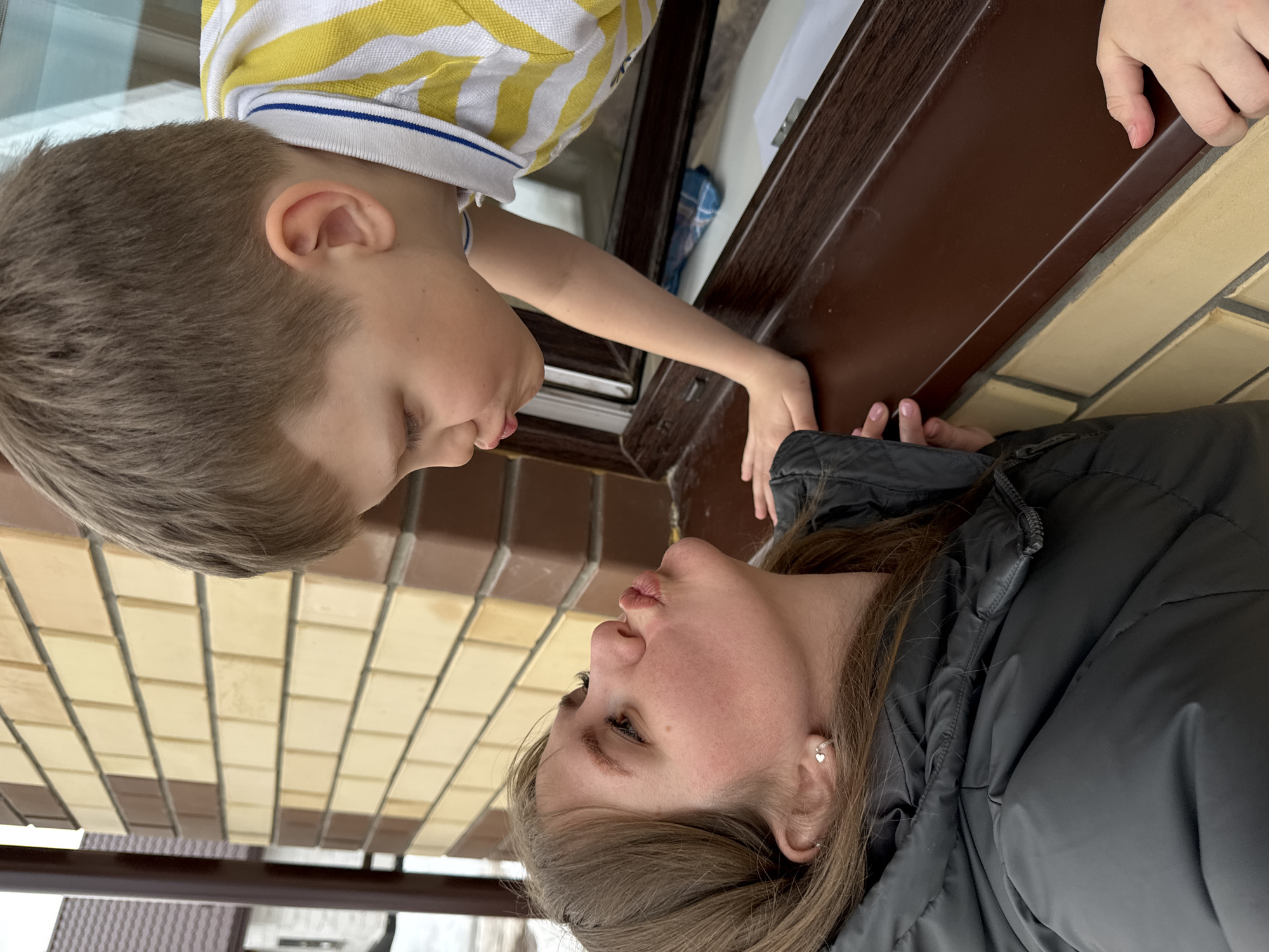 This image captures an intimate and tender moment between an adult female and a young boy, who appear to be interacting across a horizontal barrier such as a window sill or a ledge. The perspective is slightly angled, looking upwards towards the boy from the woman's side.
The young boy is positioned in the upper part of the frame, leaning down. He has short, light brown hair. He is wearing a white polo shirt with distinctive horizontal yellow and blue stripes. His face is shown in profile, looking down towards the woman, with his lips slightly puckered, suggesting he might be about to give a kiss or make a playful "kissy face." His expression appears soft and affectionate. His left arm and hand are visible, resting gently on the dark brown ledge.
The adult female is in the lower part of the frame, looking up towards the boy. She has medium-length brown hair. She is dressed in a dark, possibly black, puffer jacket, which suggests cooler weather. Her face is also in profile, mirroring the boy's, with her lips similarly puckered as if returning a kiss or engaging in a playful exchange. A small, silver heart-shaped earring is visible in her right ear. Her expression conveys warmth and affection. Her hands are also visible near the ledge, close to the boy's hands.
Between them is a dark brown, flat surface, likely a window sill or a protective ledge, which separates their physical spaces but facilitates their close interaction. The surface appears sturdy and clean.
The background behind the woman consists of a patterned wall made of light-colored bricks. The bricks are arranged in a grid pattern, with some sections appearing slightly darker than others, creating a subtle, regular texture. This brickwork suggests an outdoor setting or the exterior of a building. The background behind the boy, visible through what appears to be a window above the ledge, is lighter and blurry, indicating an interior space or a less defined outdoor area.
The overall mood of the image is affectionate and playful, capturing a sweet moment of connection between the two individuals. The lighting is soft and even, suggesting daylight without harsh shadows. No text is visible in the image.