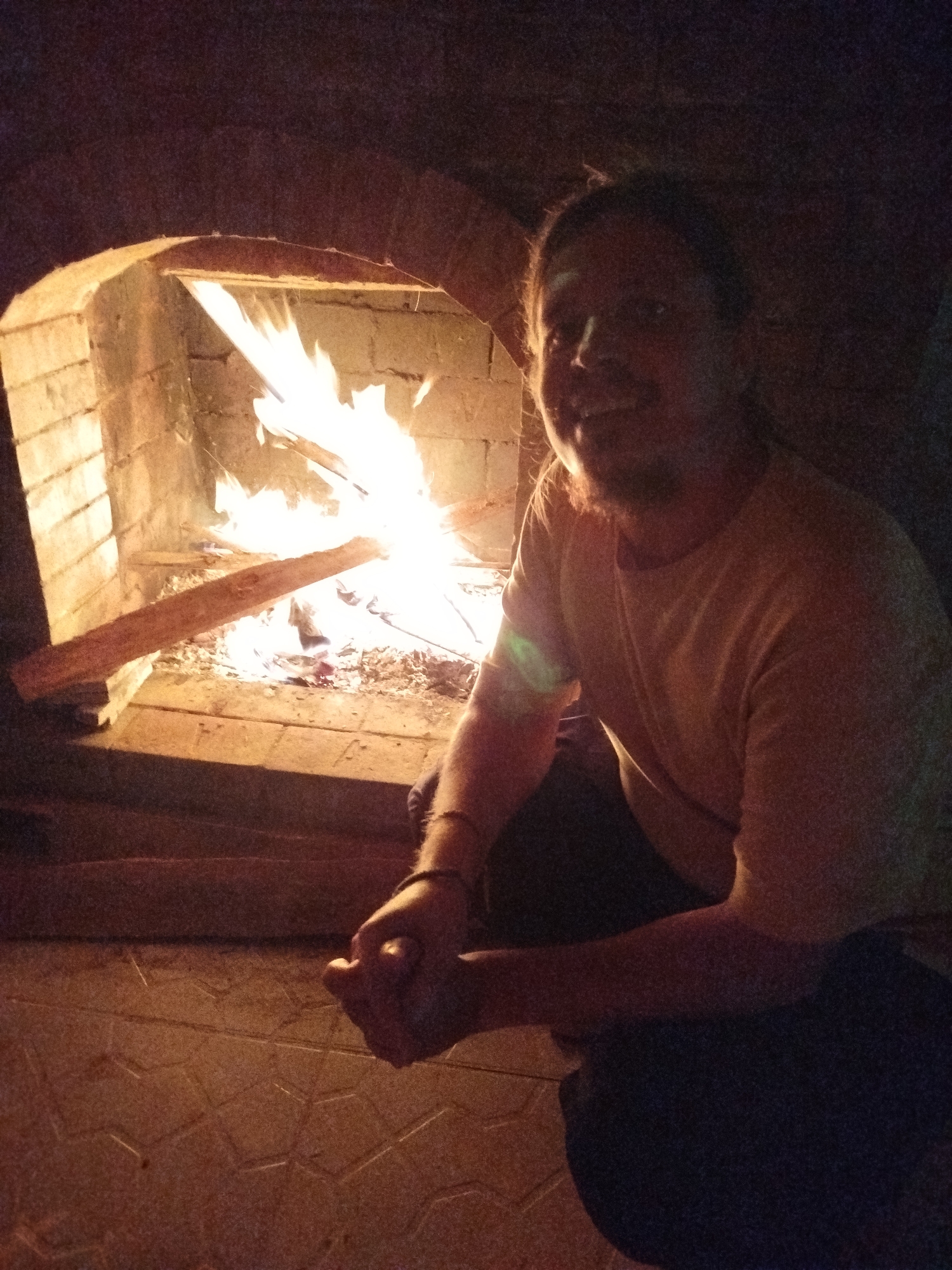A man is crouched in front of a brick fireplace with a fire burning inside. The fireplace is recessed into the brick wall. The man is wearing a light-colored t-shirt and dark pants. His hands are clasped in front of him. The floor appears to be tiled. No further information about location is available.