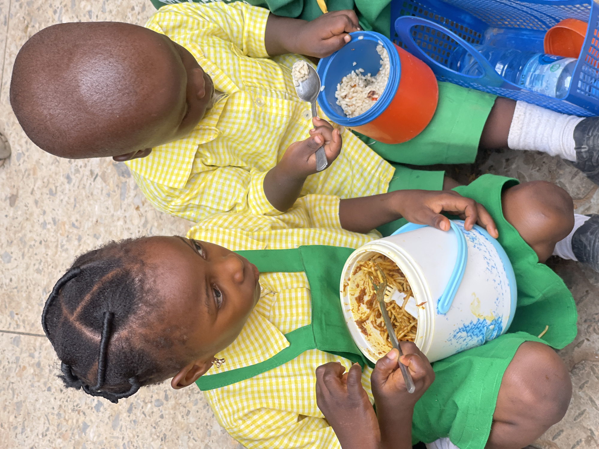 Two young Black children, dressed in yellow and white checkered shirts and green aprons, are eating meals outdoors on a paved surface in Akwanga, Nigeria. The child on the left, a girl with braided hair, is holding a fork and a plastic container filled with spaghetti. The child on the right, a boy with a shaved head, is using a spoon to eat rice from an orange container. A blue plastic basket containing a water bottle and an orange lid is beside them. The ground is a textured grey pavement.