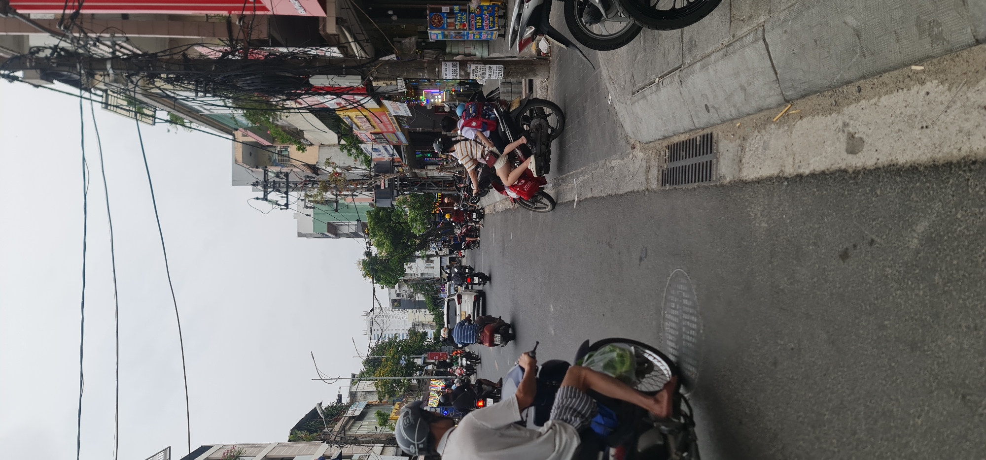 The image depicts a busy street in what appears to be Đà Nẵng, Vietnam. There is heavy traffic consisting mainly of scooters and motorbikes. The street is lined with buildings and shops on both sides. A dense network of electrical wires hangs overhead. Many people can be seen on the scooters, and they are wearing various types of casual clothing. The weather seems overcast.