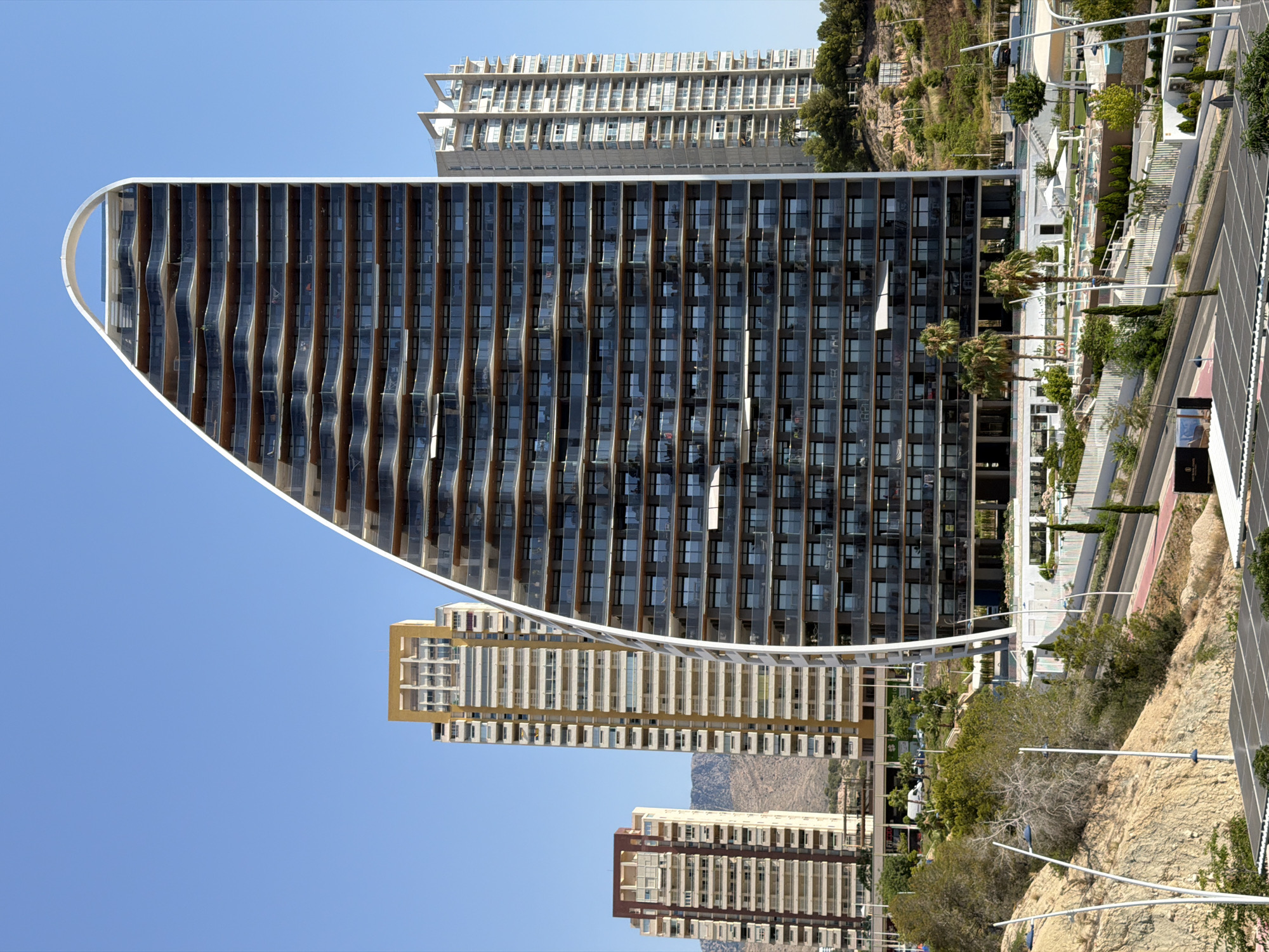 This is a photograph of a modern, multi-story building with a distinctive curved facade, made of dark glass and wood-toned panels. The building is located in a coastal city, likely Benidorm, Spain, given the clear blue sky and the presence of other high-rise buildings in the background, typical of the Mediterranean resort town.
The building's design features a unique wavy pattern on its exterior, created by the arrangement of its balconies and windows. In front of this main structure, there's another tall building with a lighter, yellowish facade and a more traditional rectangular design. Further in the background, another very tall building with a beige, tiered structure is visible, as well as some rocky, vegetated hillsides.
The scene appears to be a sunny day, with bright, clear skies suggesting it's daytime, likely late morning or afternoon given the lighting. There is no discernible activity or event taking place; the image focuses on the architecture and the urban landscape. The setting appears to be a developed area with some landscaping, including palm trees, and what looks like a swimming pool area or public space with white umbrellas. There's also a road or pathway visible in the foreground with markings. No people are clearly visible in the frame. There is no visible text in the image.